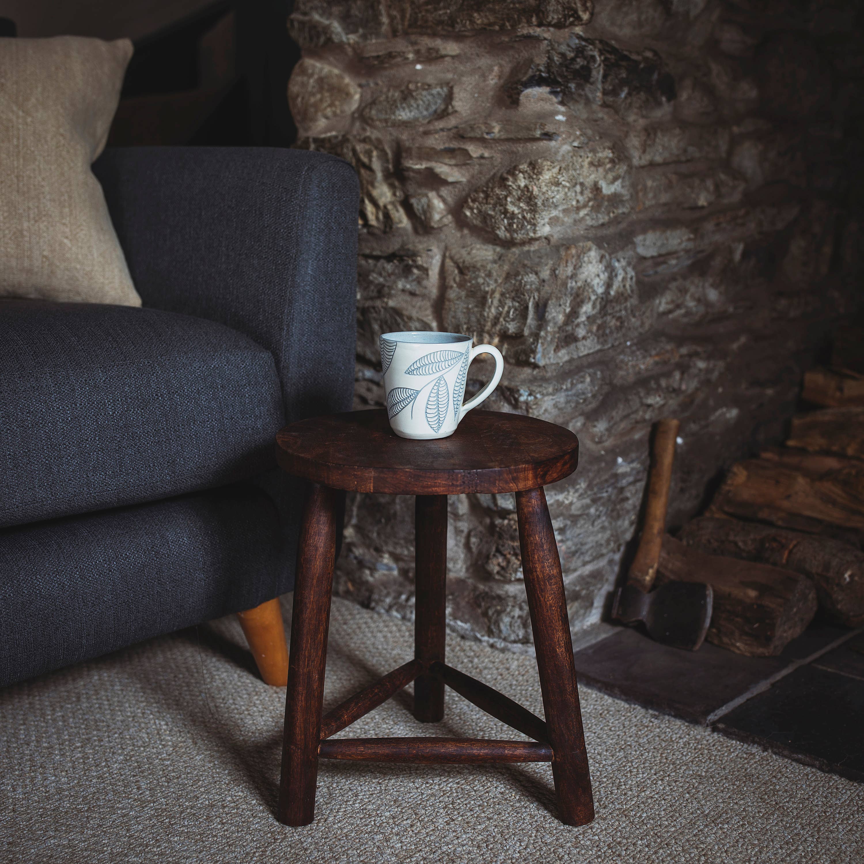 Interior shot of Mango stool by stone fireplace and navy armchair . Mug on top of the stool.Lovely dark mango wood, ethically sourced in India.