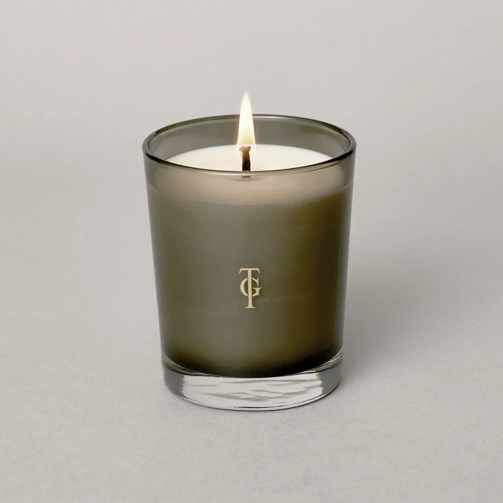 Studio shot of lit candle - smoked glass holder has True Grace Logo on glass. Candle is white. Beautiful , luxurious gift.