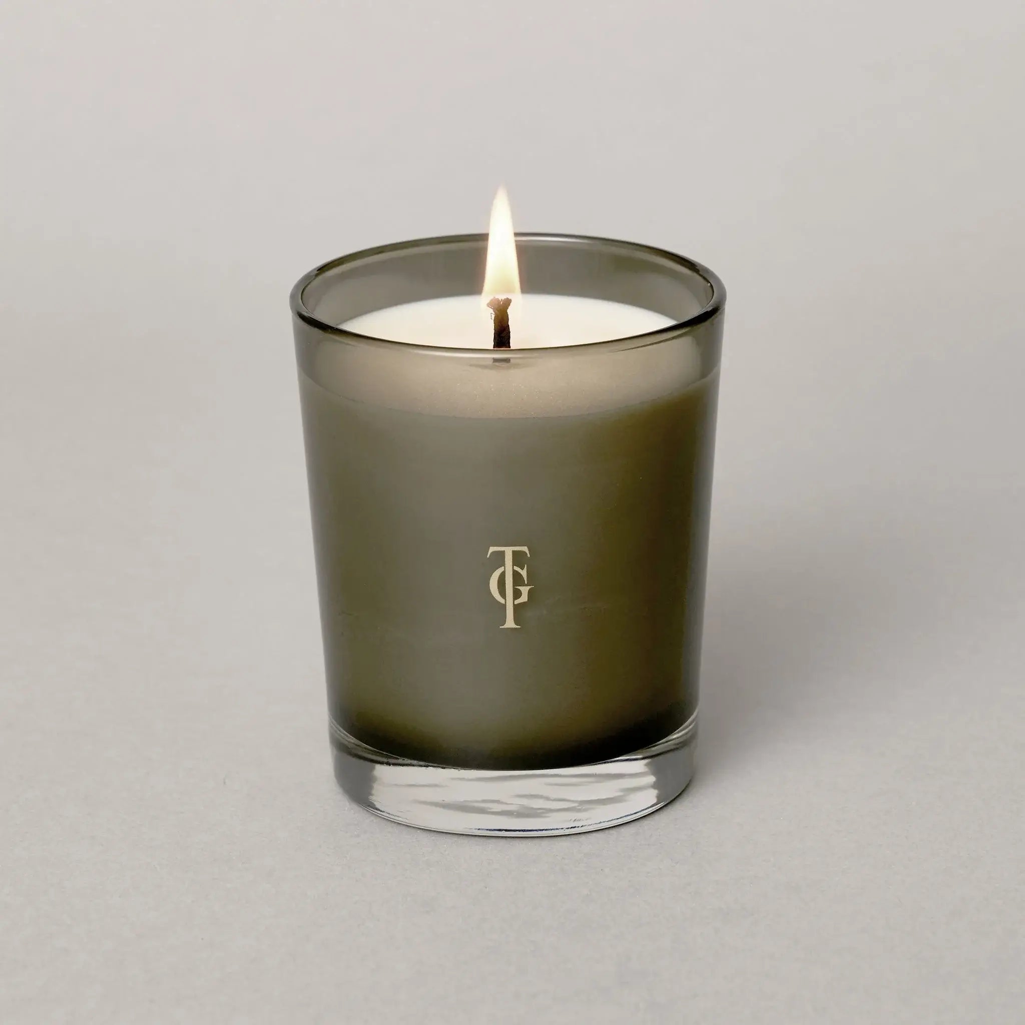 Studio shot of lit candle - smoked glass holder has True Grace Logo on glass. Candle is white. Beautiful , luxurious gift.