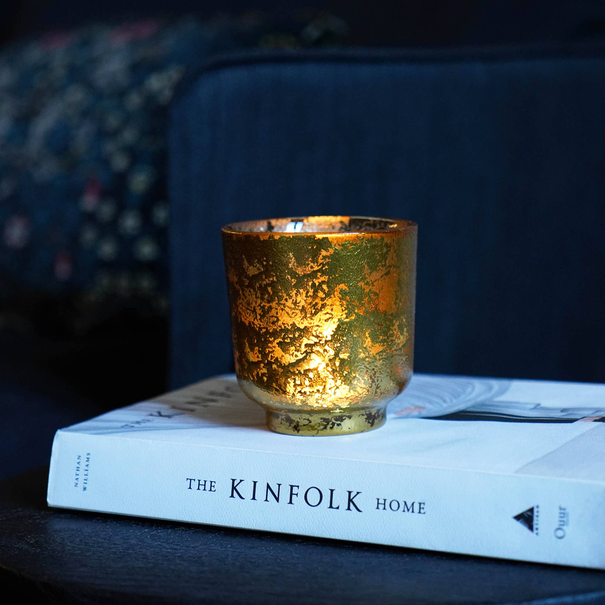 Golden Glass Tealight holder on top of a book . The spine of the book is showing the text ' The Kinfolk Home" Interior shot with a blue sofa in background.
