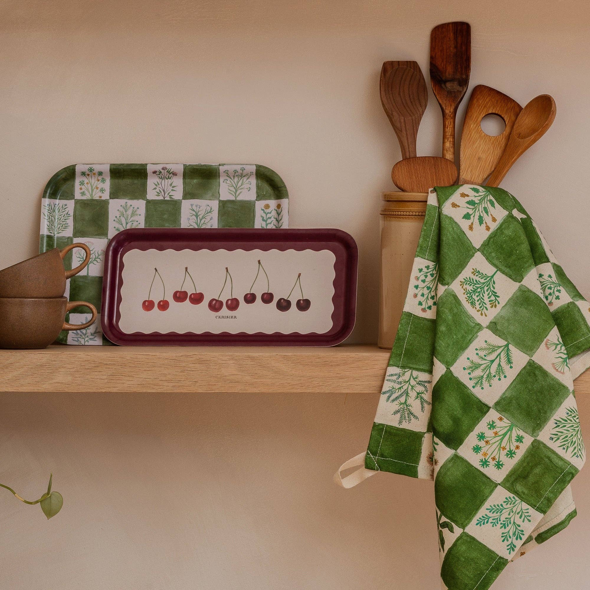 Kitchen shelf with some mugs and trays , utensils jar and tea towel which is opened out and hanging from a utensil.