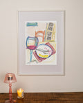 Framed artwork of a dining scene with a glass, plate, and chair on a wall above a table with a lamp and candle.