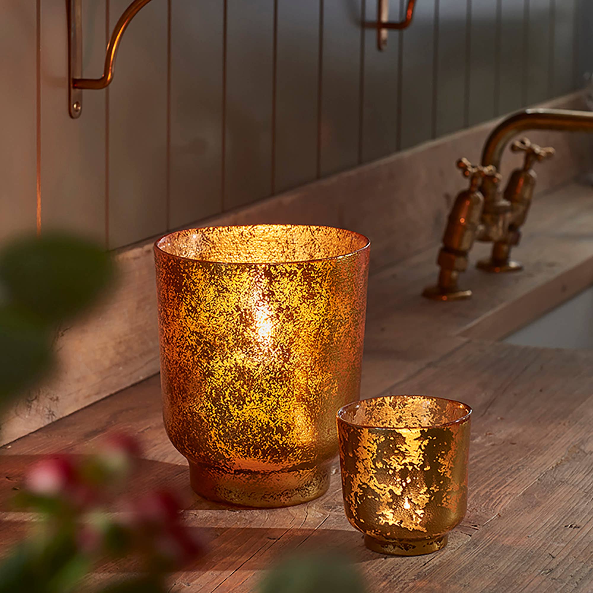 Large and small copper tea light holders on wooden kitchen surface by sink. Both are internally glowing.