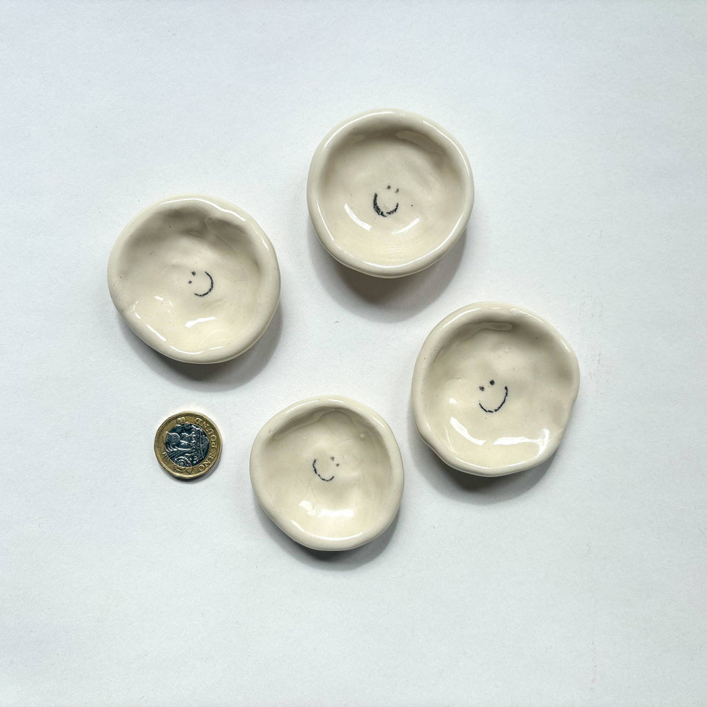 Four  handmade ceramic dishes with smiley face design  are shown beside a pound coin for scale. Handmade unique gifts.