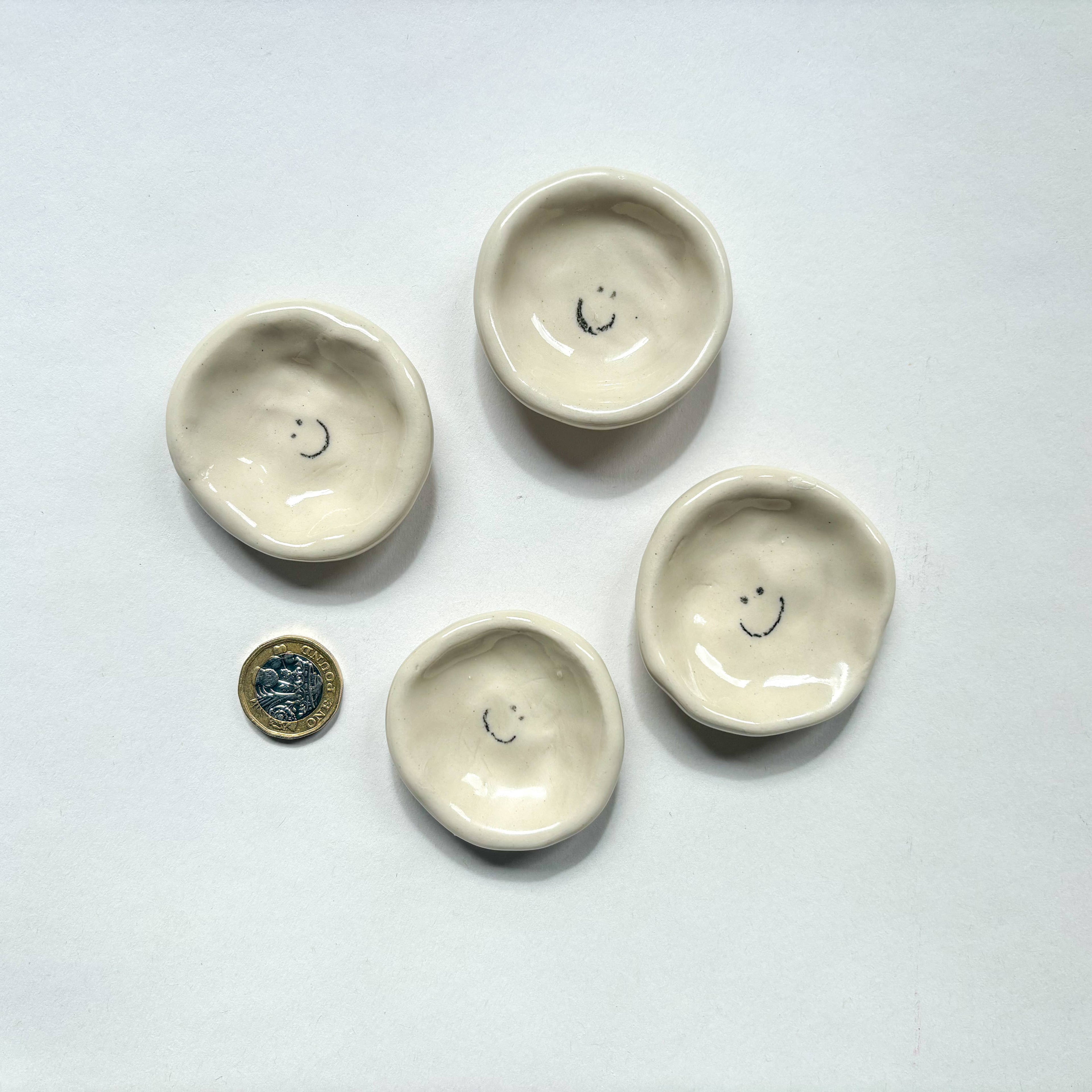Four  handmade ceramic dishes with smiley face design  are shown beside a pound coin for scale. Handmade unique gifts.
