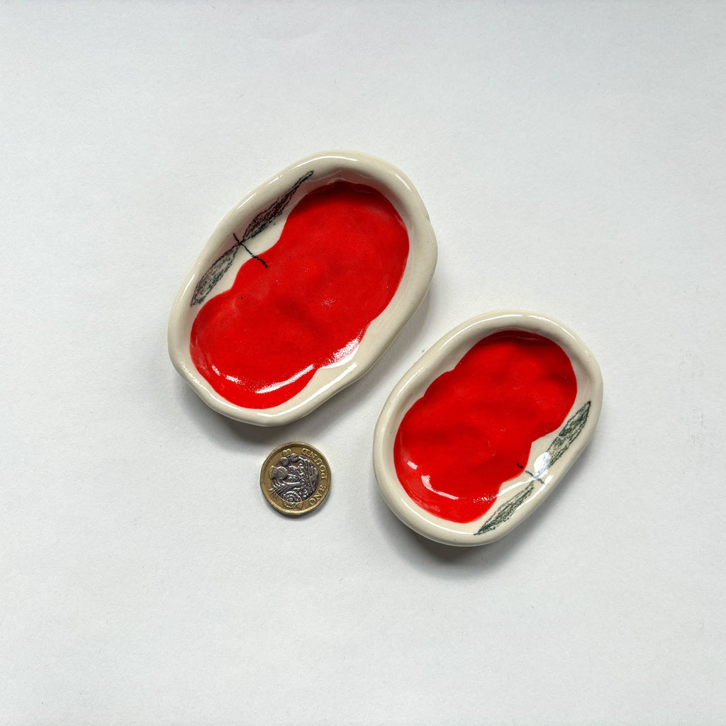 Two  oval handmade ceramic dishes with a red apple design  are shown beside a pound coin for scale. Handmade unique gifts.