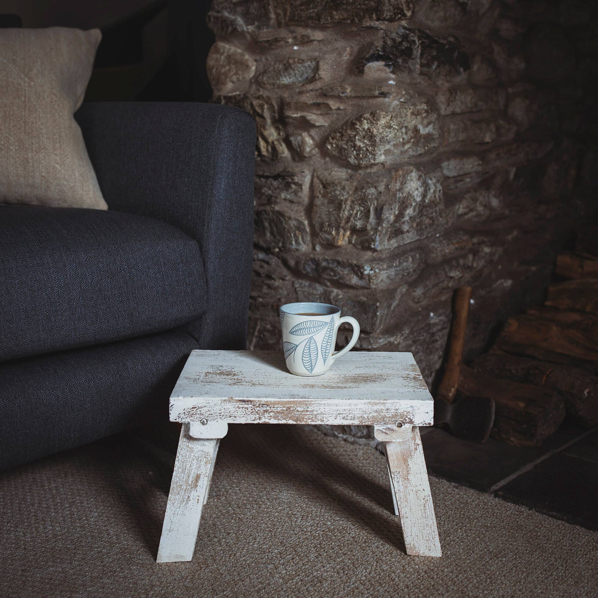 Interior shot by stone fireplace and navy armchair of white stool with a mug on the top.
Folding whitewashed stool - perfect for resting a mug on.Made in India and ethically sourced.