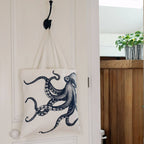 Interior shot of doorway with octopus tote bag hanging on a metal hook on white door. A wooden surface is in the background with a plant pot and green plant.