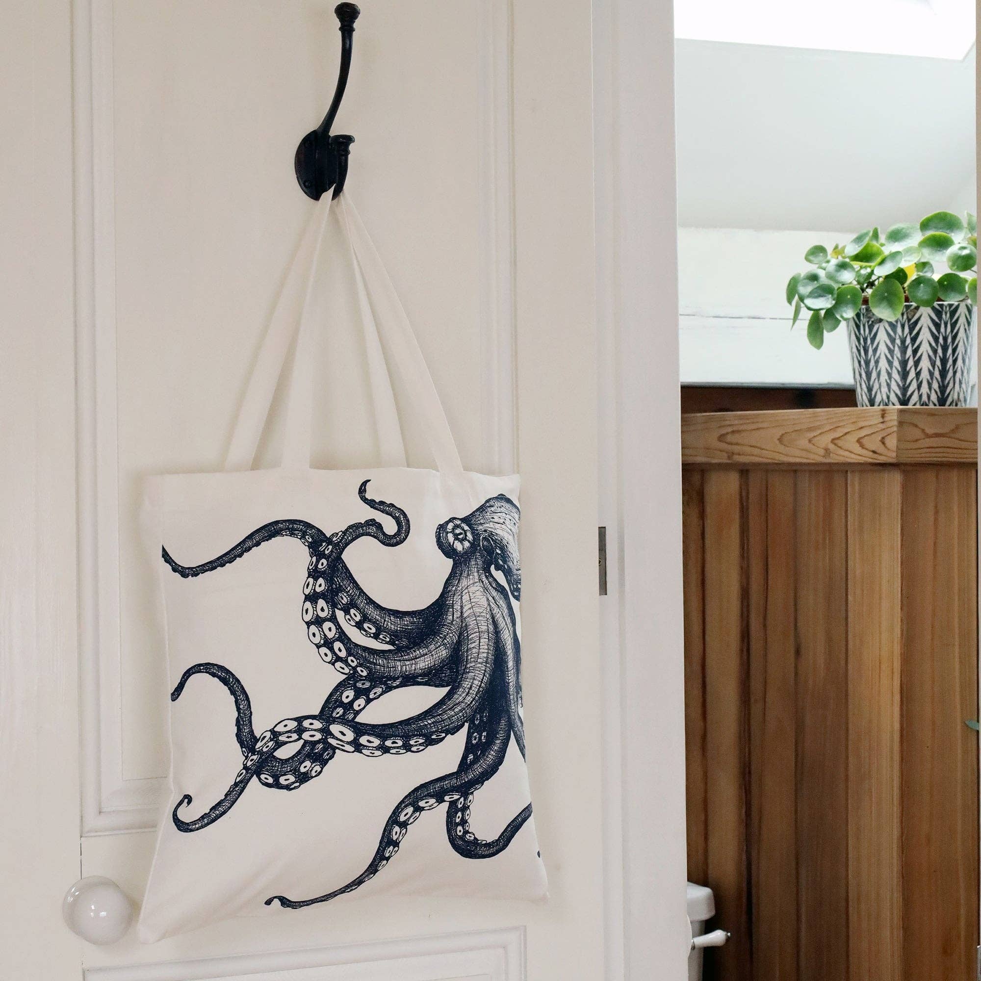 Interior shot of doorway with octopus tote bag hanging on a metal hook on white door. A wooden surface is in the background with a plant pot and green plant.