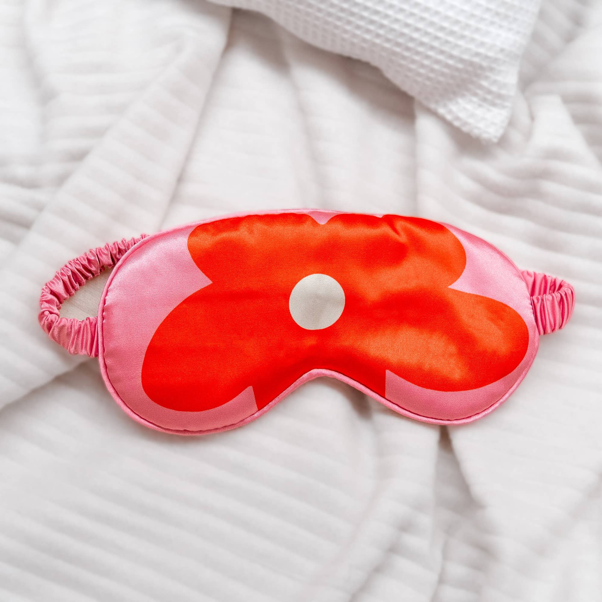 Red and pink bold flower design sleep mask with pink straps on a white textured fabric background.