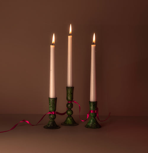 Three green verdigris candlesticks in  varying heights tied together with a silky hot pink ribbon. The candle sticks each have a lit cream candle  giving a soft dusky glow in the caramel coloured backdrop.