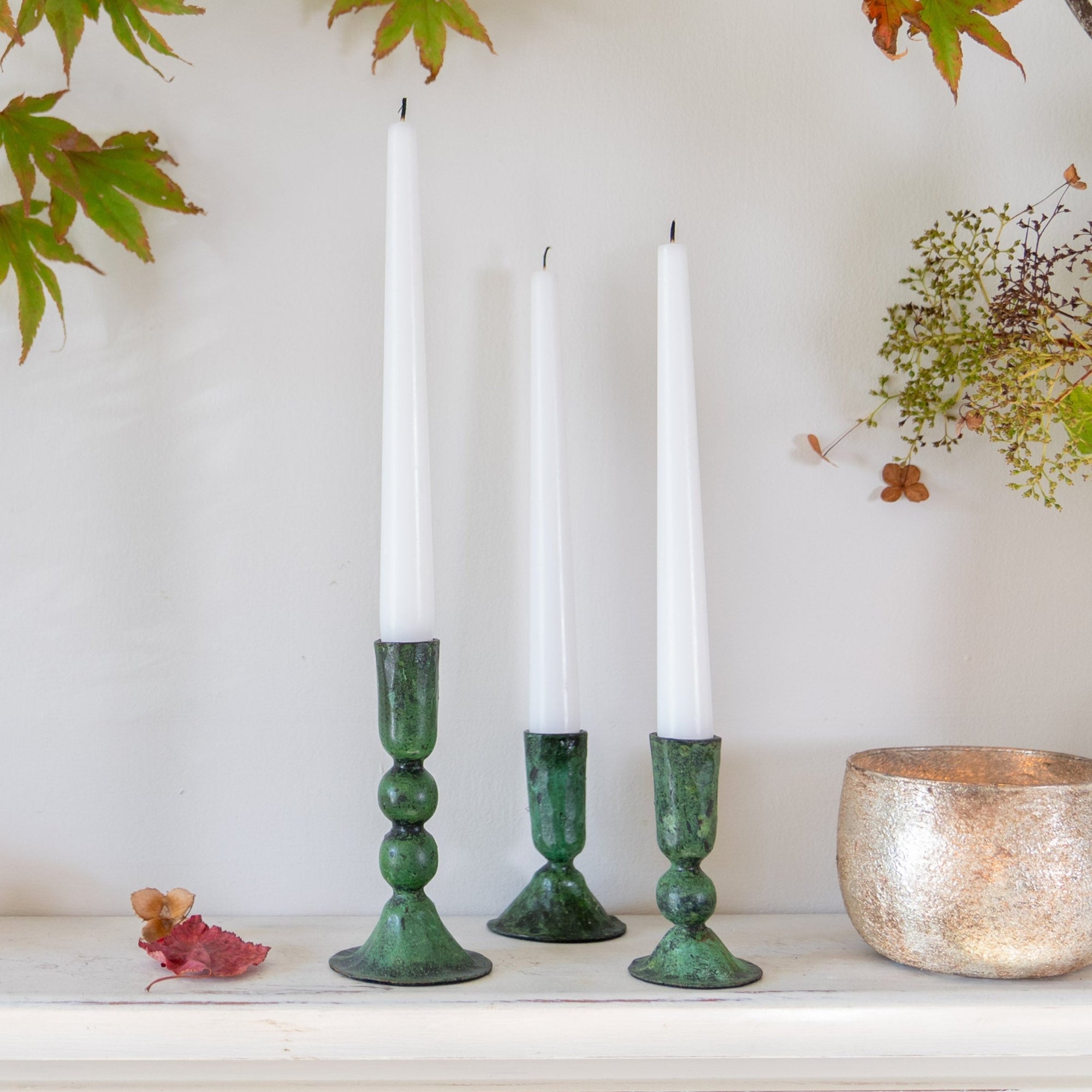 Three green verdigris candlesticks on a white mantlepiece.The white candles are unlit and some autumn leaves and a gold bowl are also in the vignette.  Handmade verdigris iron candlesticks.