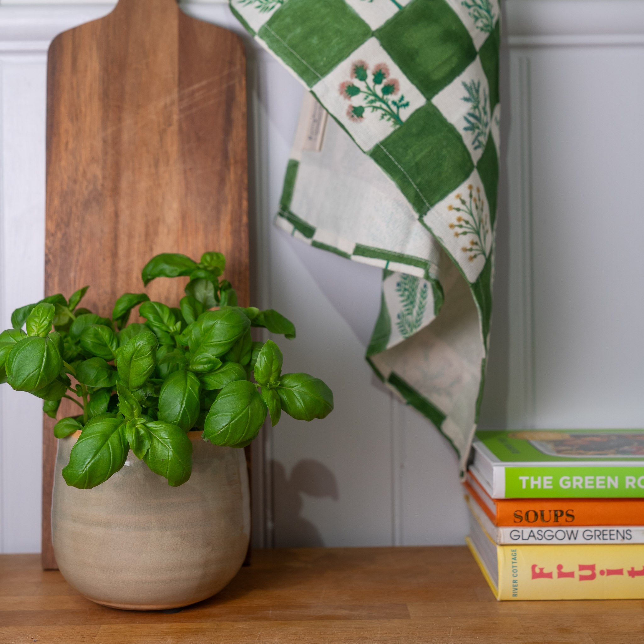 A ceramic Ivyline planter with herbs next to a green and white checkered Roomytown organic herb tea towel, all placed on a wooden surface. Chopping board and recipe books are alongside.