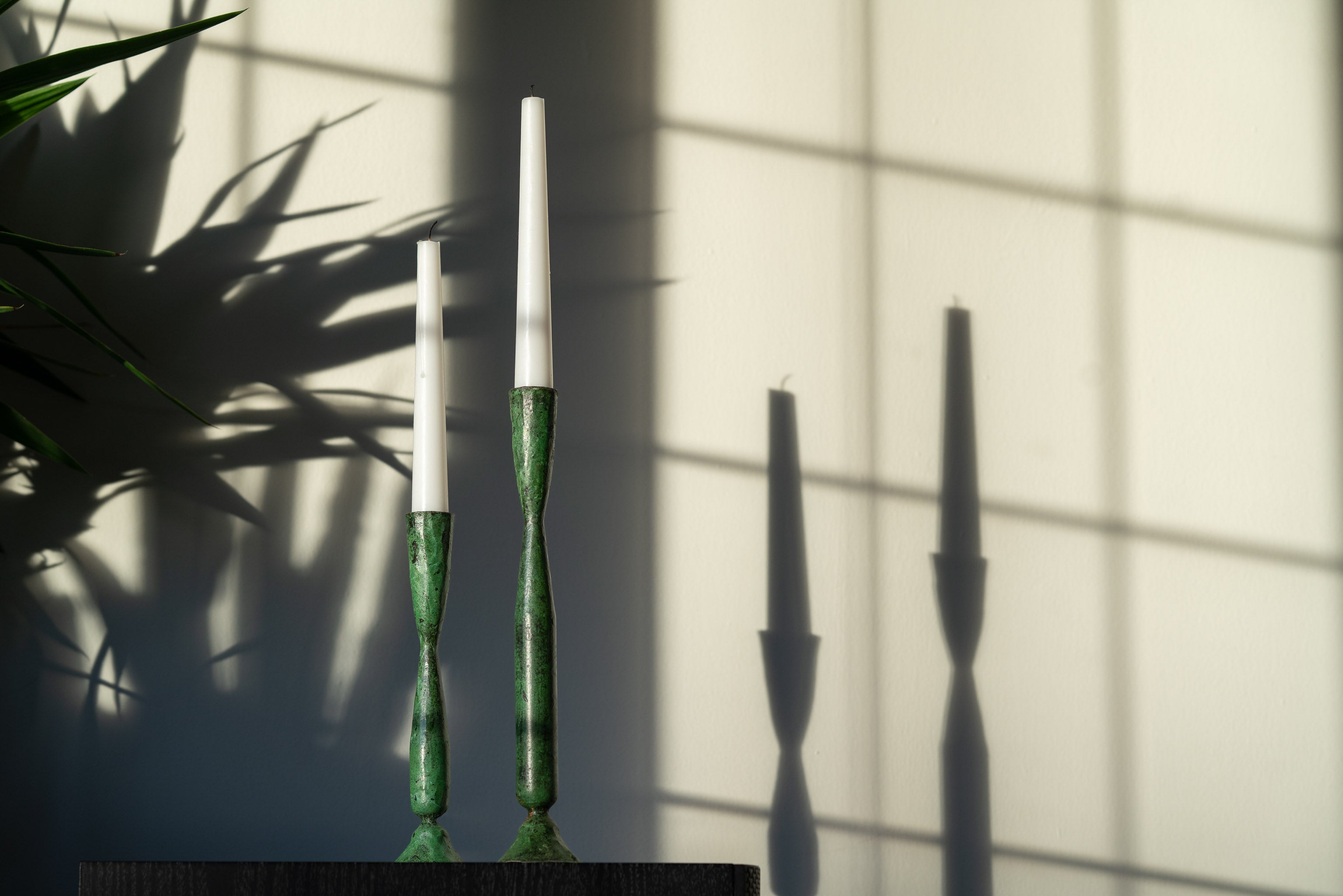 Pair of verdigris candlesticks med and large. Shot in sunlight with silhouette of  plant and shadows on background wall. Hand made Shelley tall verdigris iron candlesticks.