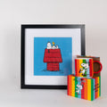A gift set featuring a framed print of Snoopy on a red dog house and a multicolored mug with Snoopy design, presented on a white background. The mug has a colourful gift box - perfect gift for a Snoopy fan!