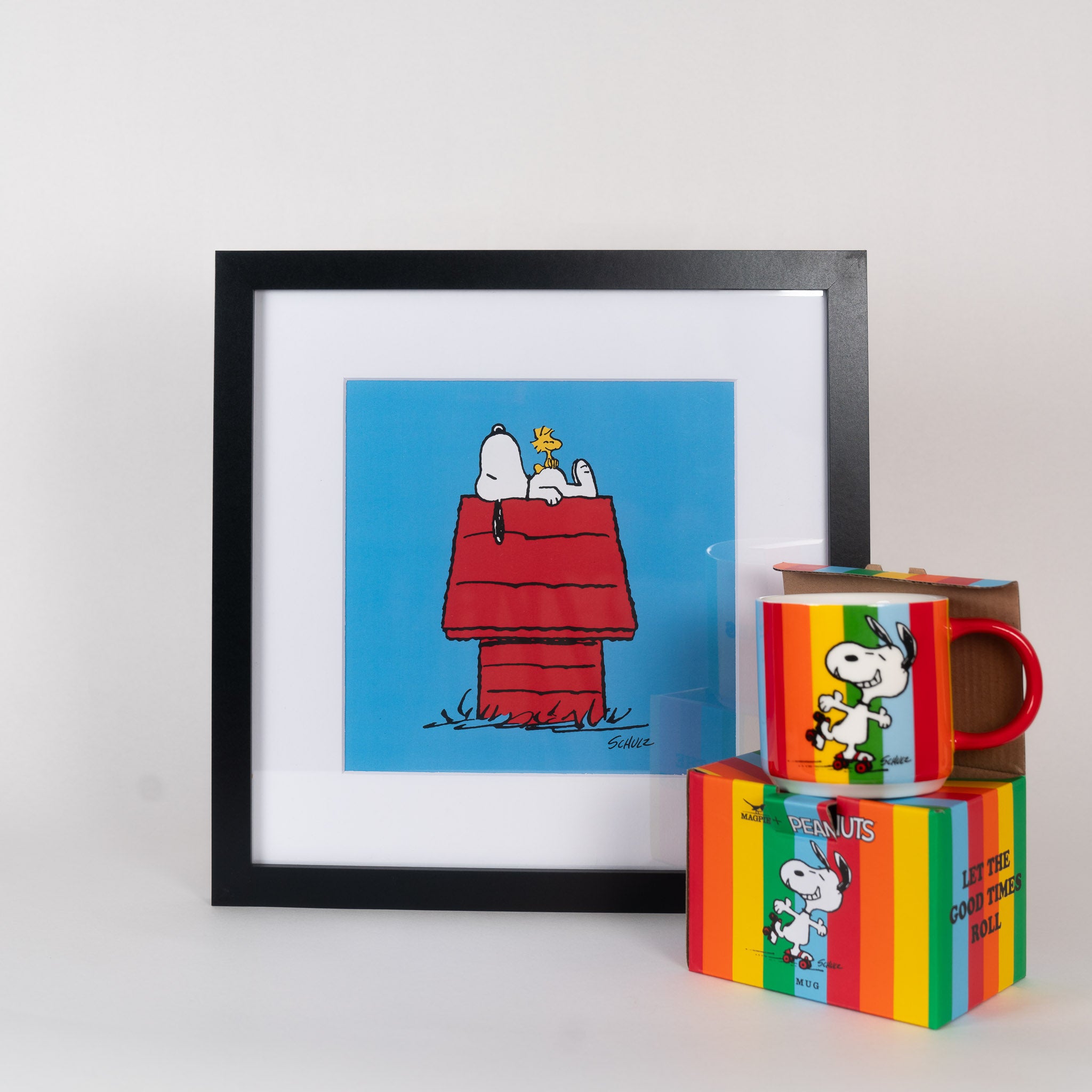 A gift set featuring a framed print of Snoopy on a red dog house and a multicolored mug with Snoopy design, presented on a white background. The mug has a colourful gift box - perfect gift for a Snoopy fan!