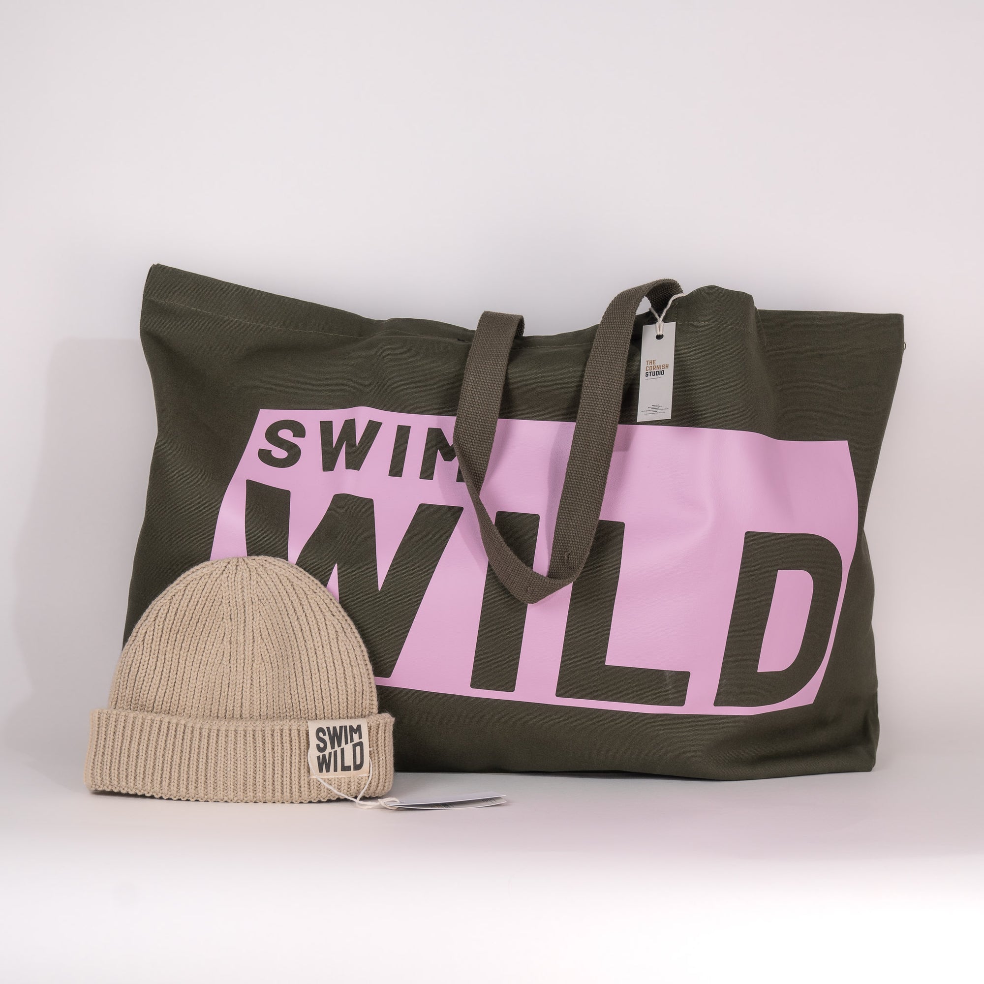 A jumbo olive cotton tote bag with 'SWIM WILD' printed in pink, accompanied by a beige beanie hat with 'SWIM WILD' text.