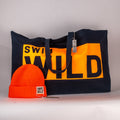 A black cotton tote bag with 'SWIM WILD' text in orange accompanied by an orange knit hat with 'SWIM WILD' band. Great gift bundle from the Cornish Studio.