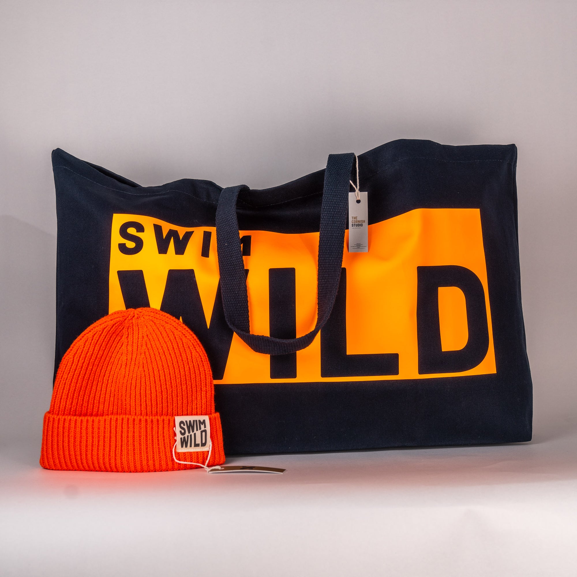 A black cotton tote bag with 'SWIM WILD' text in orange accompanied by an orange knit hat with 'SWIM WILD' band. Great gift bundle from the Cornish Studio.