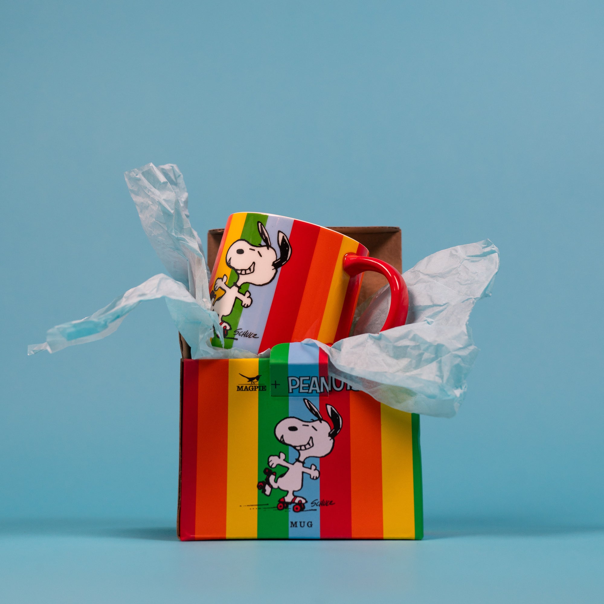 Colourful Peanuts gift box open with tissue  spilling out and Peanuts Good Times mug against a blue background.