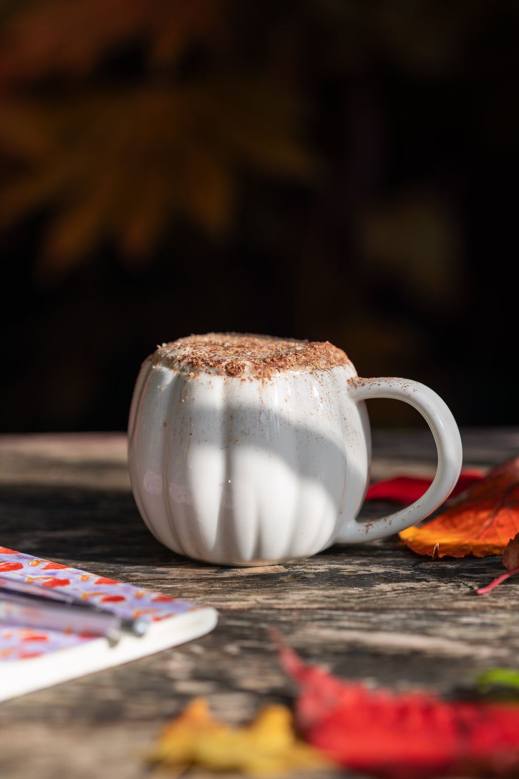 Pumpkin shaped mug on outdoor table with autumn leaves.Filled with cinnamon topped drink.