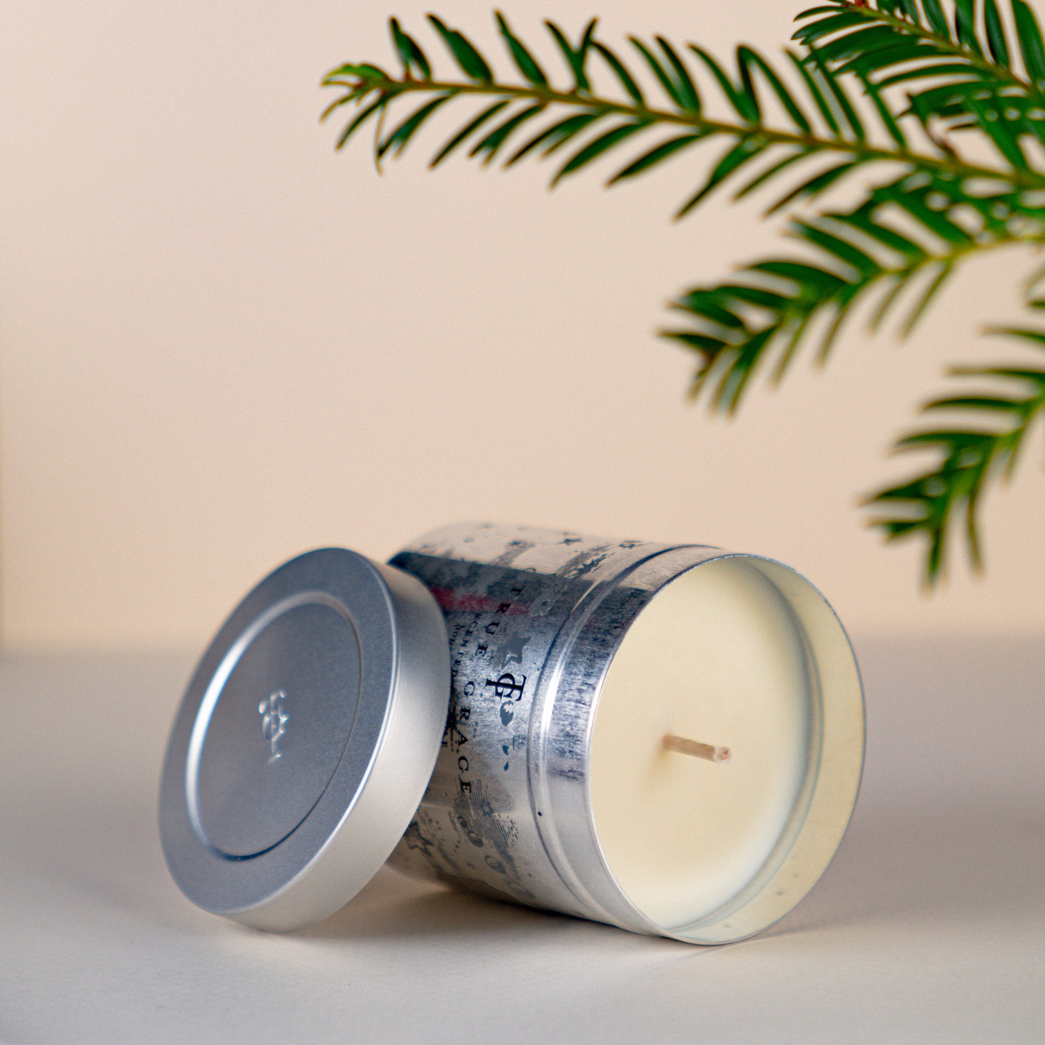 True Grace Manor Christmas Tin Candle. Cream background with some pine foliage. Candle is on side with lid off showing creamy wax and wick.