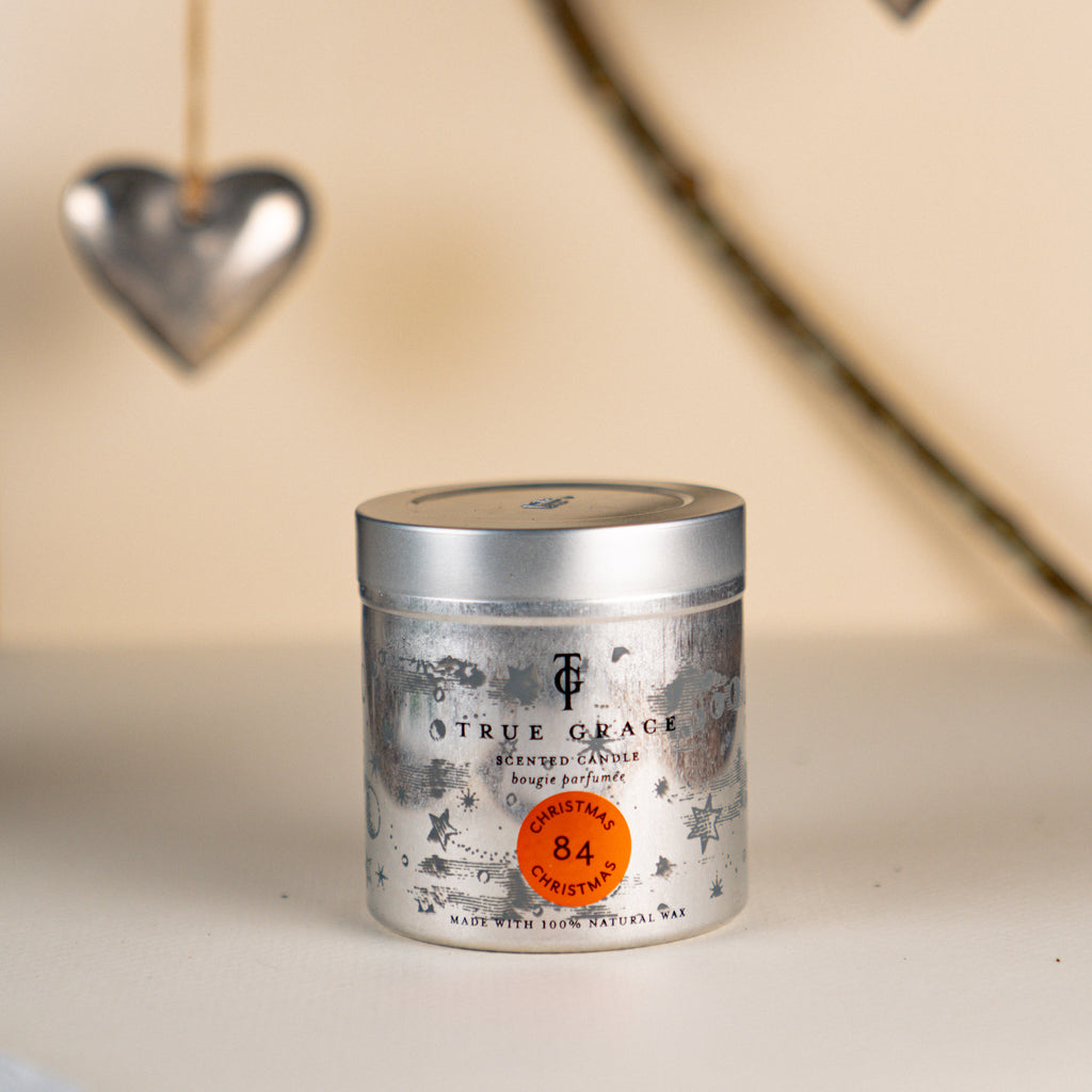 True Grace Christmas Christmas Candle in a Tin number 84 on a  cream background with a branch and hanging silver heart.