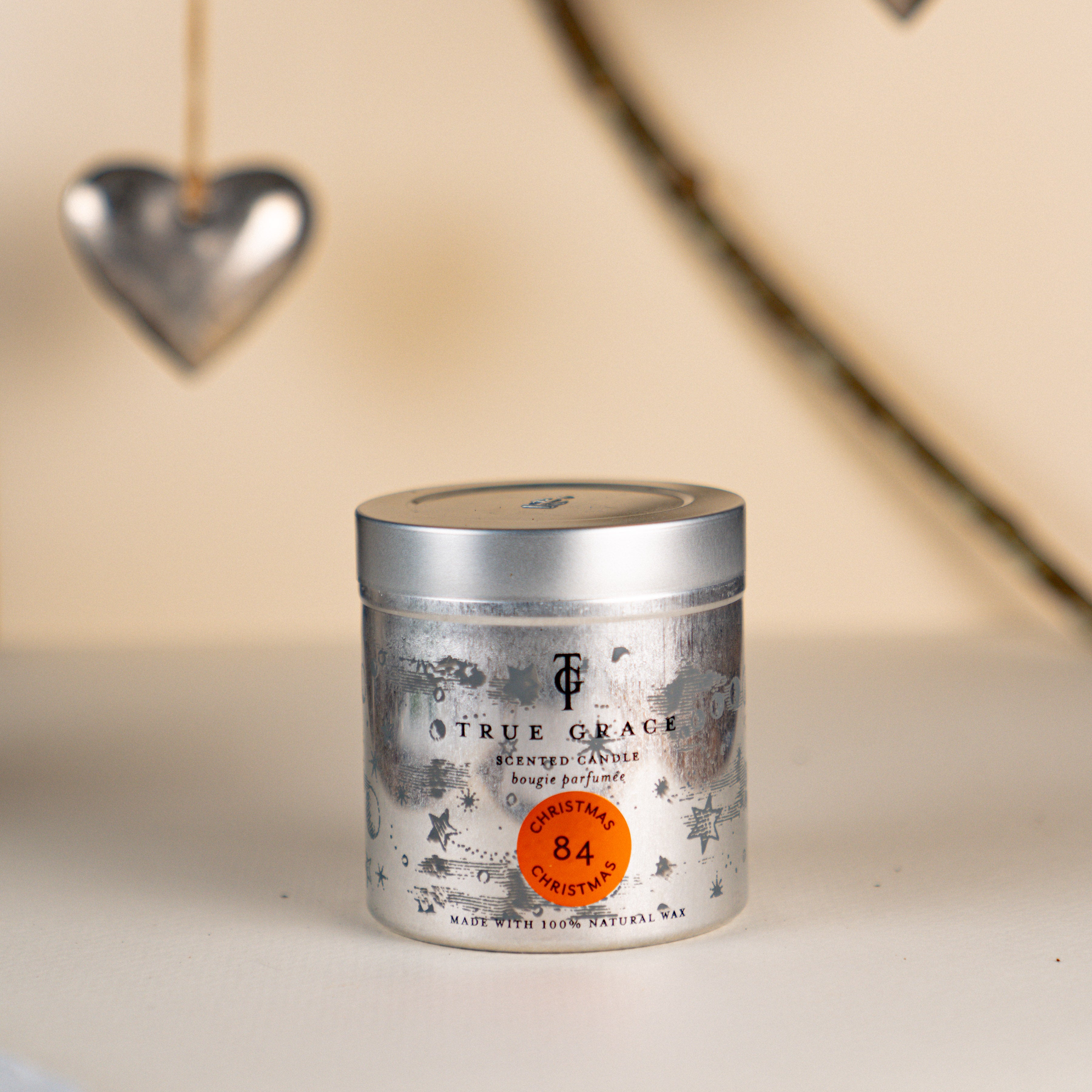True Grace Christmas Christmas Candle in a Tin number 84 on a  cream background with a branch and hanging silver heart.