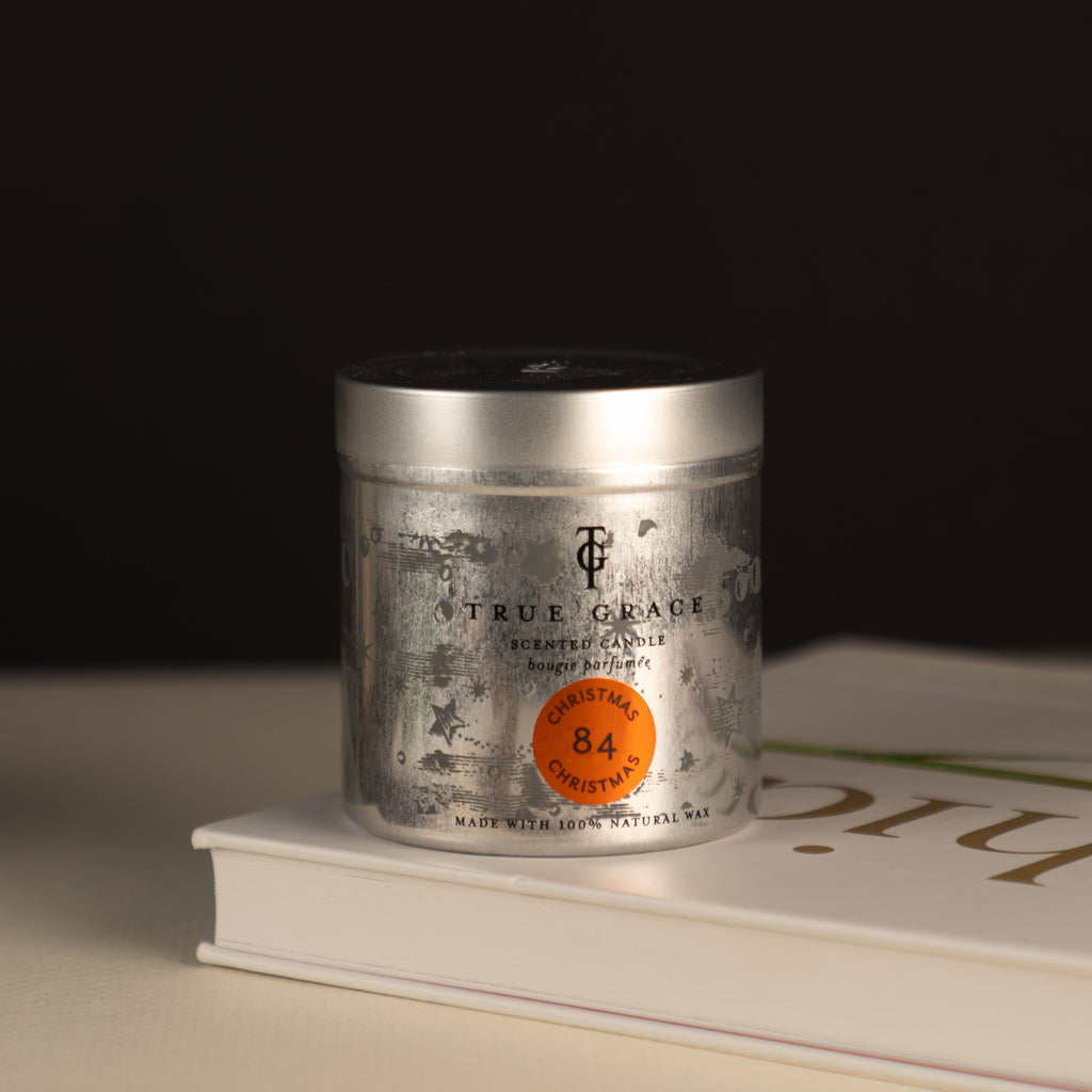 True Grace Christmas Christmas Candle in a Tin number 84 on a black background and placed on top of a coffee table style book.