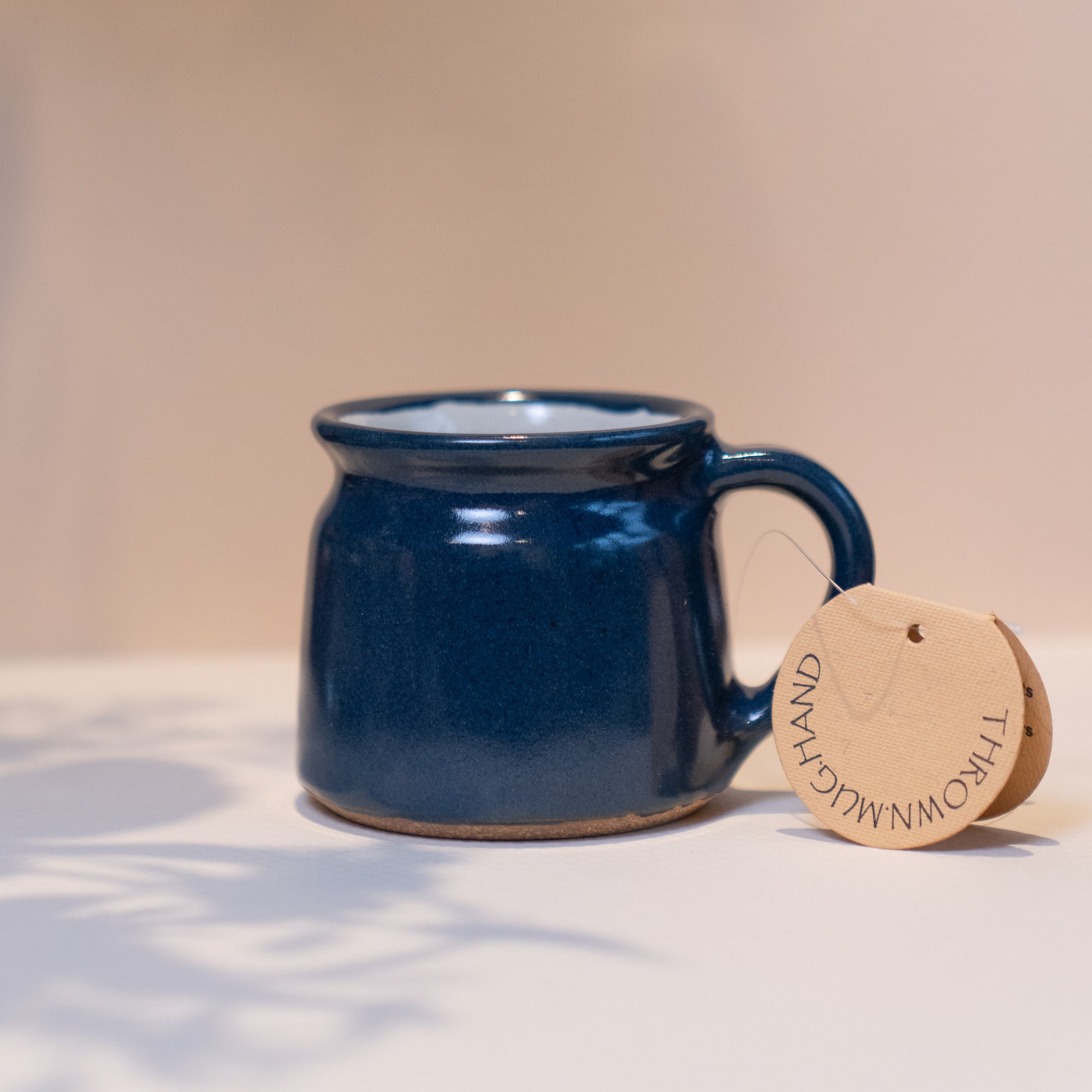 Hand made navy blue ceramic mug on a light warm coloured backround. Mug label reads  'THROWN.MUG.HAND' 
