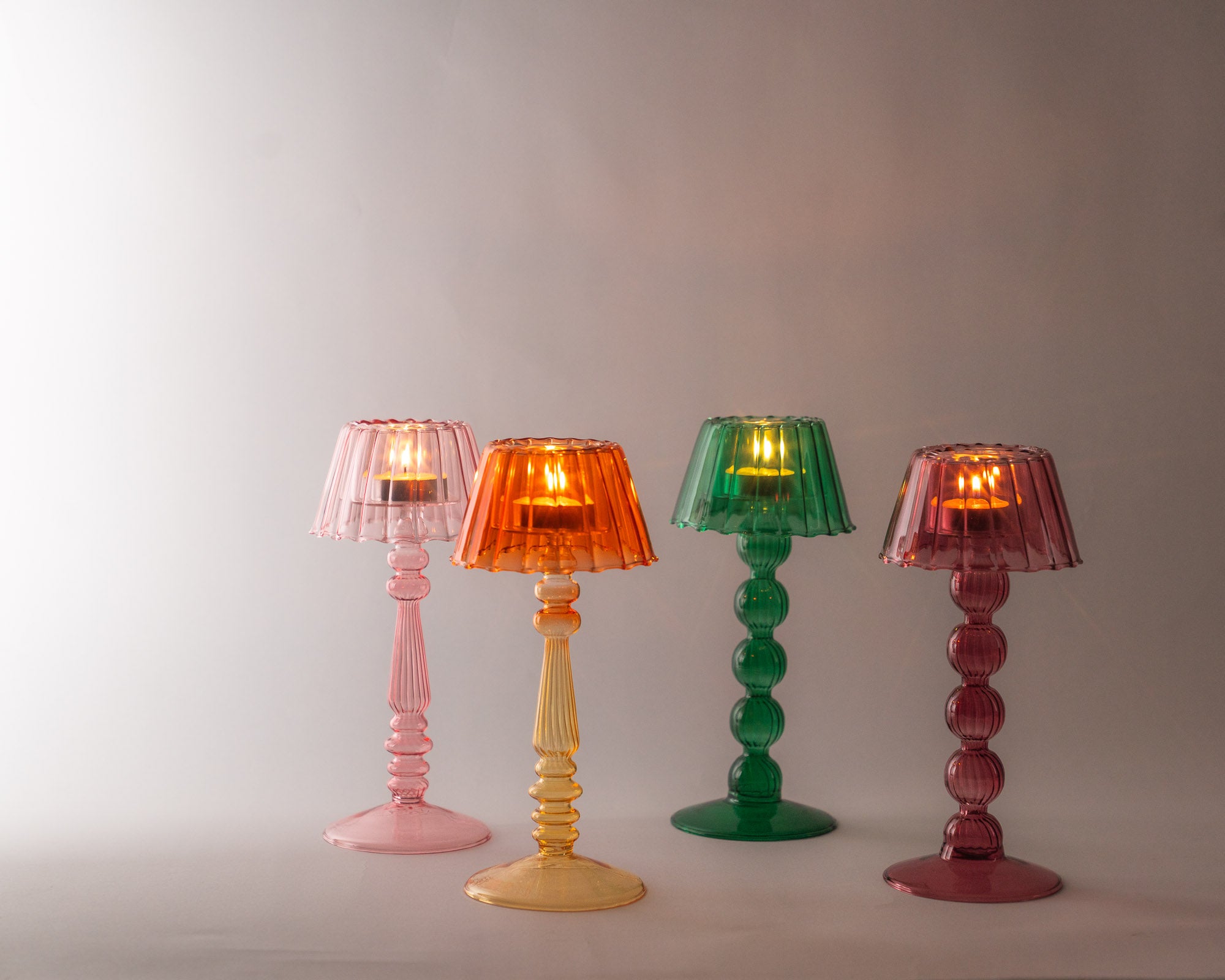 Four colourful glass lamps on a plain background - shown with tea lights lit and all are softly glowing.