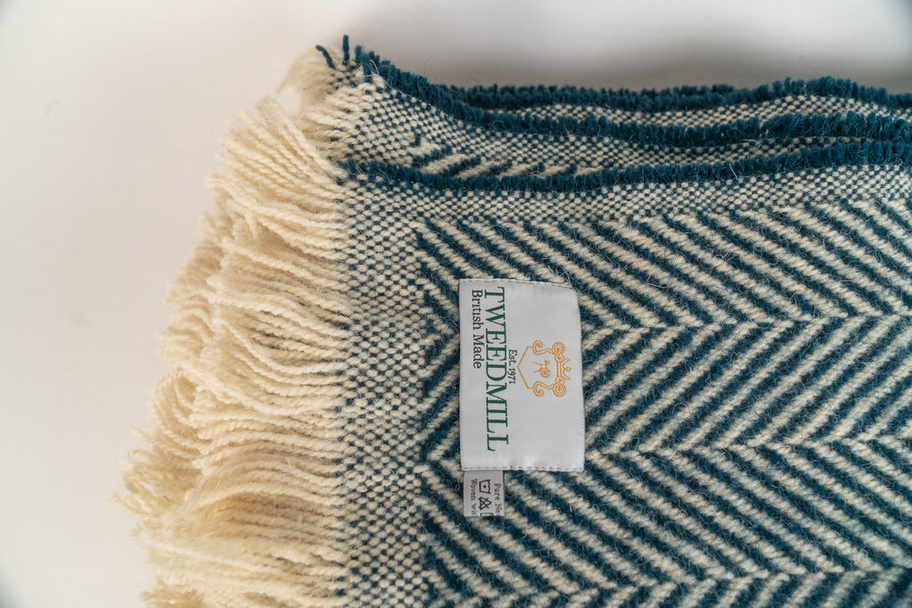 Section of a 100% recycled wool blanket with a herringbone pattern and Tweedmill label on a white background. The rug is Pacific Blue and has cream fringing.