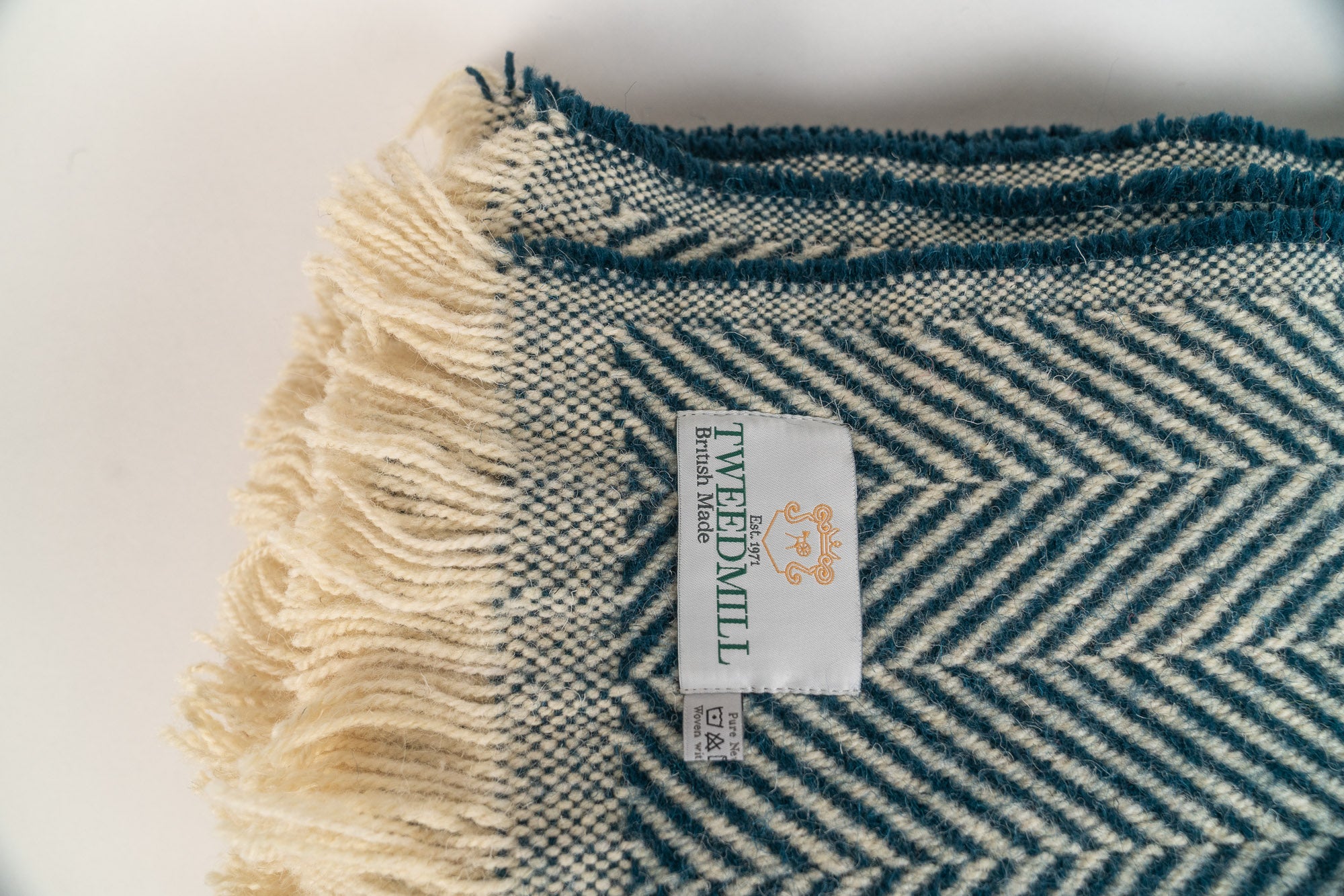 Section of a 100% recycled wool blanket with a herringbone pattern and Tweedmill label on a white background. The rug is Pacific Blue and has cream fringing.