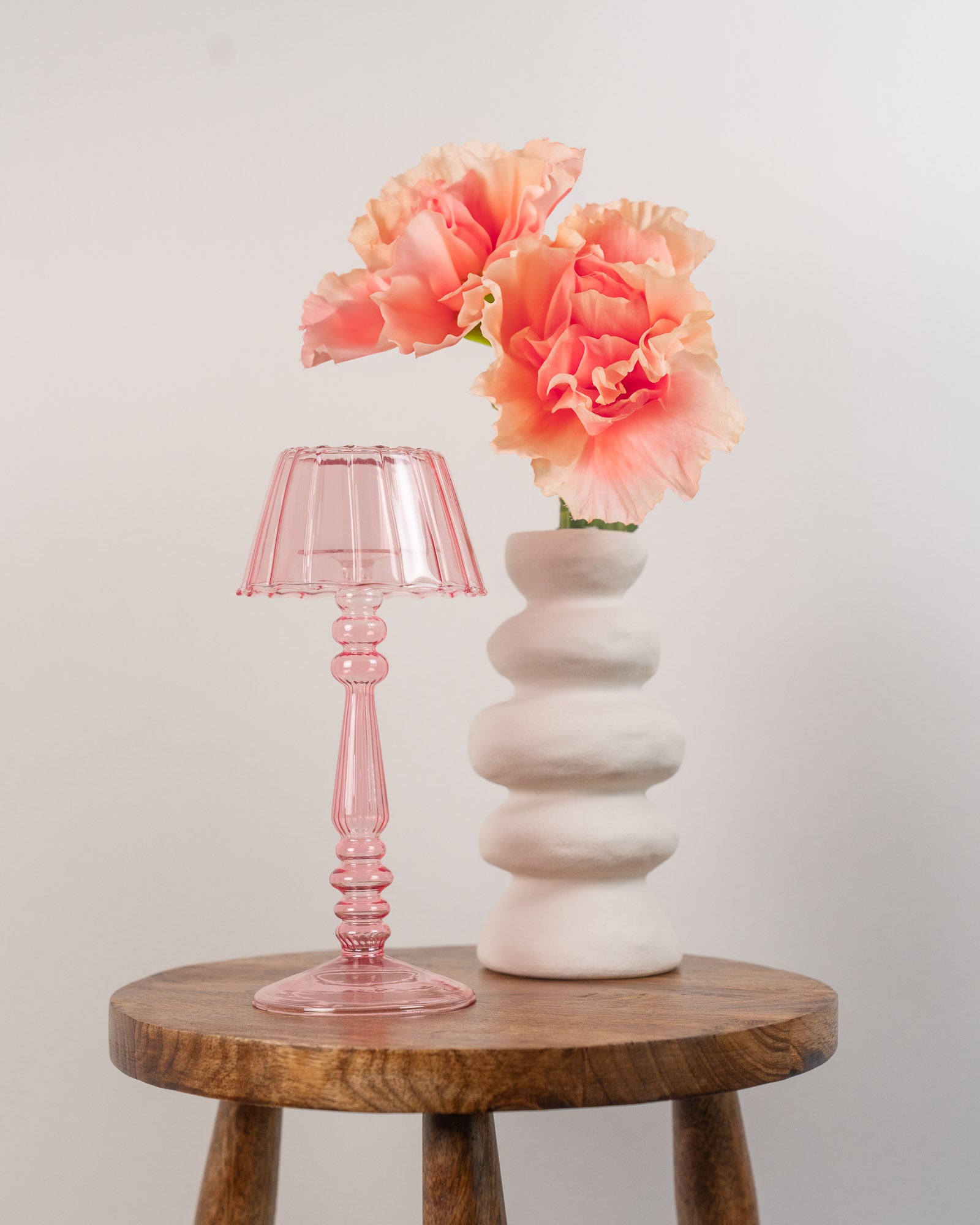 Pink glass tea light holder and white vase with flowers on a wooden stool against a white background