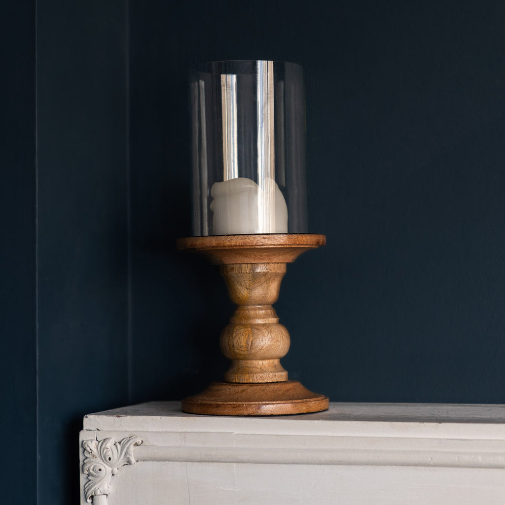 Wood and glass pillar candle holder with unlit cream candle, on white mantelpiece against dark blue wall.