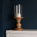 Wood and glass pillar candle holder with unlit cream candle, on white mantelpiece against dark blue wall.