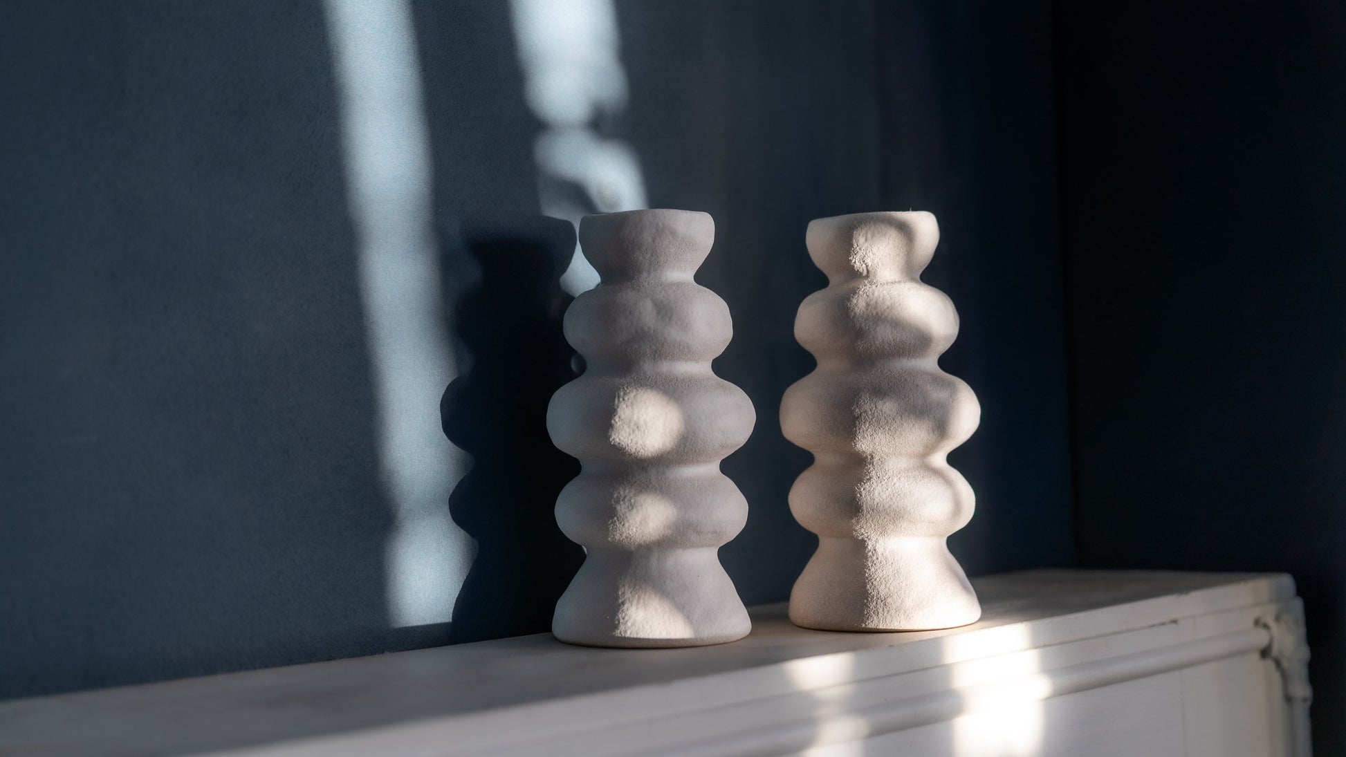 two white vases on a white shelf infront of a dark blue wall. Abigail Ahern designed vases with wavey curves and a stone texture. Vases are lit by 2 rays of sun casting strong shadows of their wavey silhouettes.