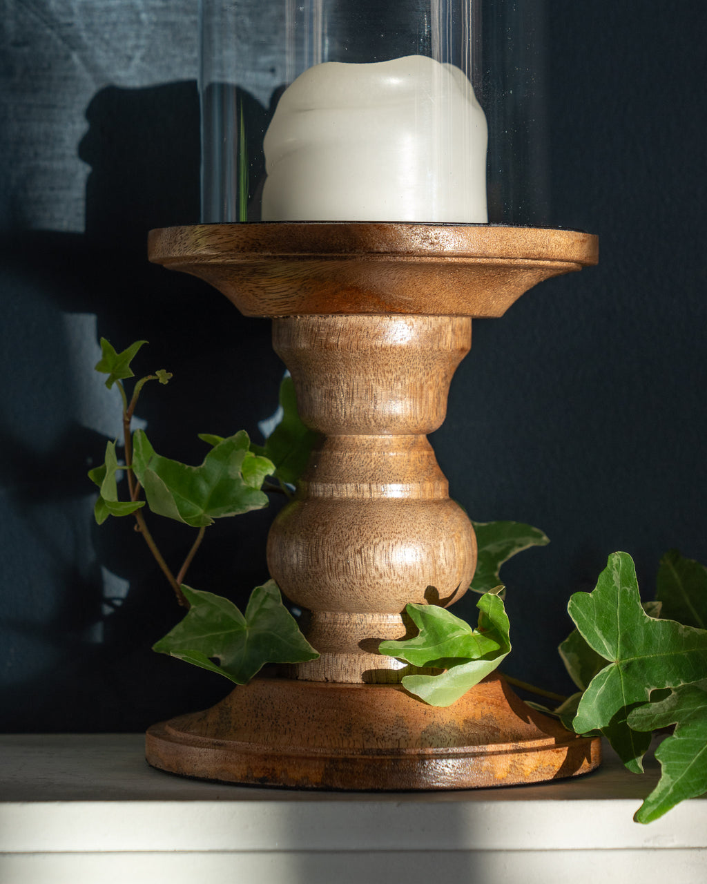 Close up of wood and glass pillar candle holder with unlit cream candle, on white mantelpiece against dark blue wall, ivy curled around base.