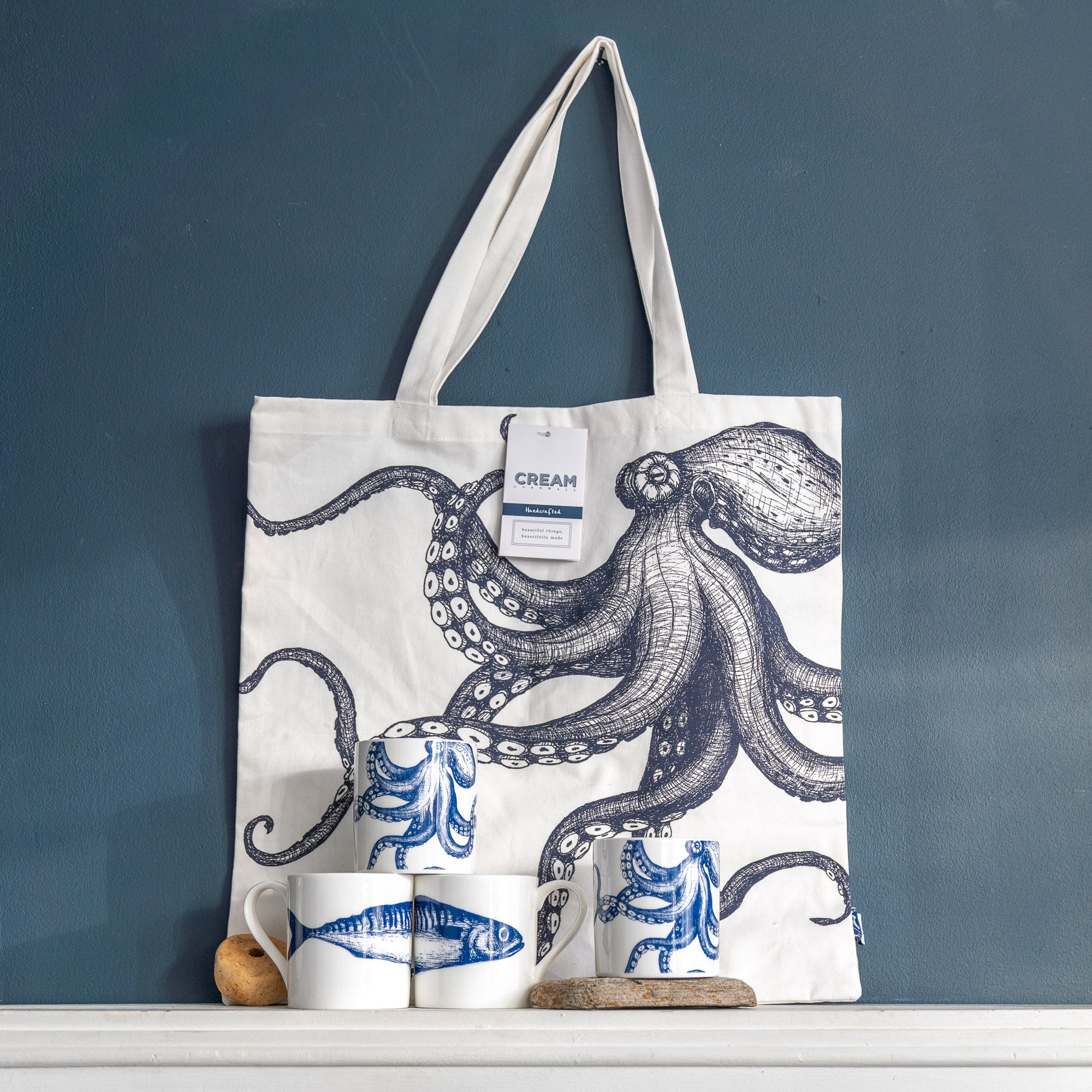 Tote bag with octopus design on a shelf against a blue wall. Beside it are  four mugs, two are octopus design and two are mackerel fish deign. Cream Cornwall gifting ideas.