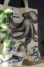 Tote bag with octopus design and mugs some green fronds of a plant are in the foreground.