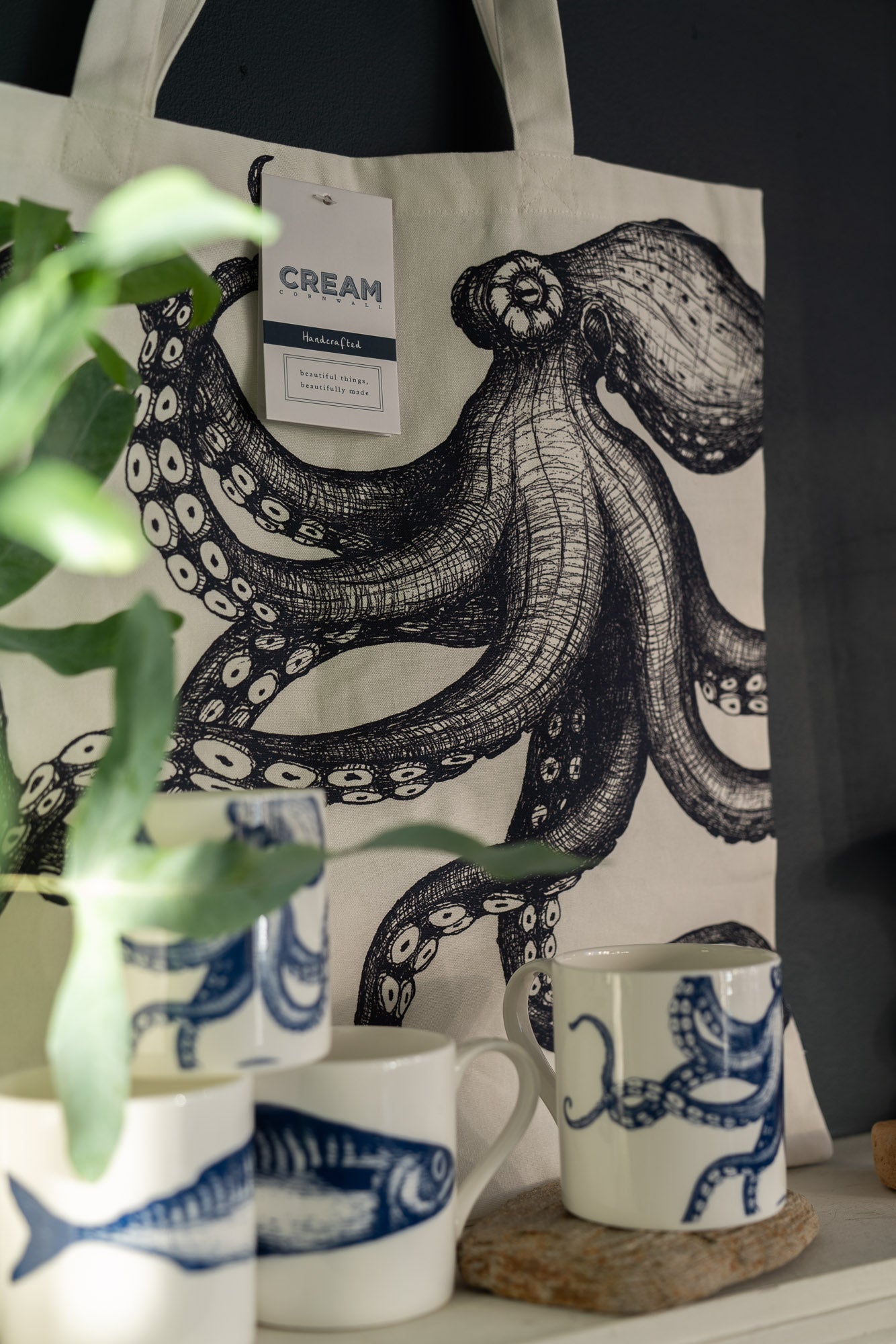 Tote bag with octopus design and mugs some green fronds of a plant are in the foreground.