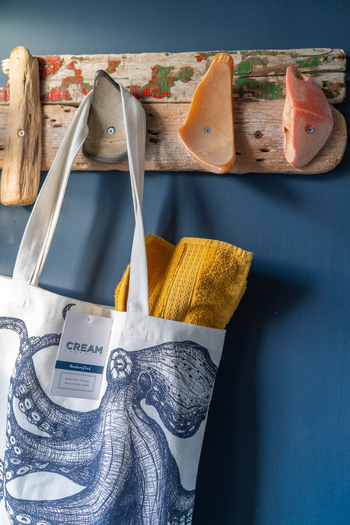 Tote bag with octopus design and 'Cream Cornwall' label . Hanging from  driftwood  on a hook made from repurposed bits of broken beach buoys. The tote bag has a yellow rolled towel peeping out the top.