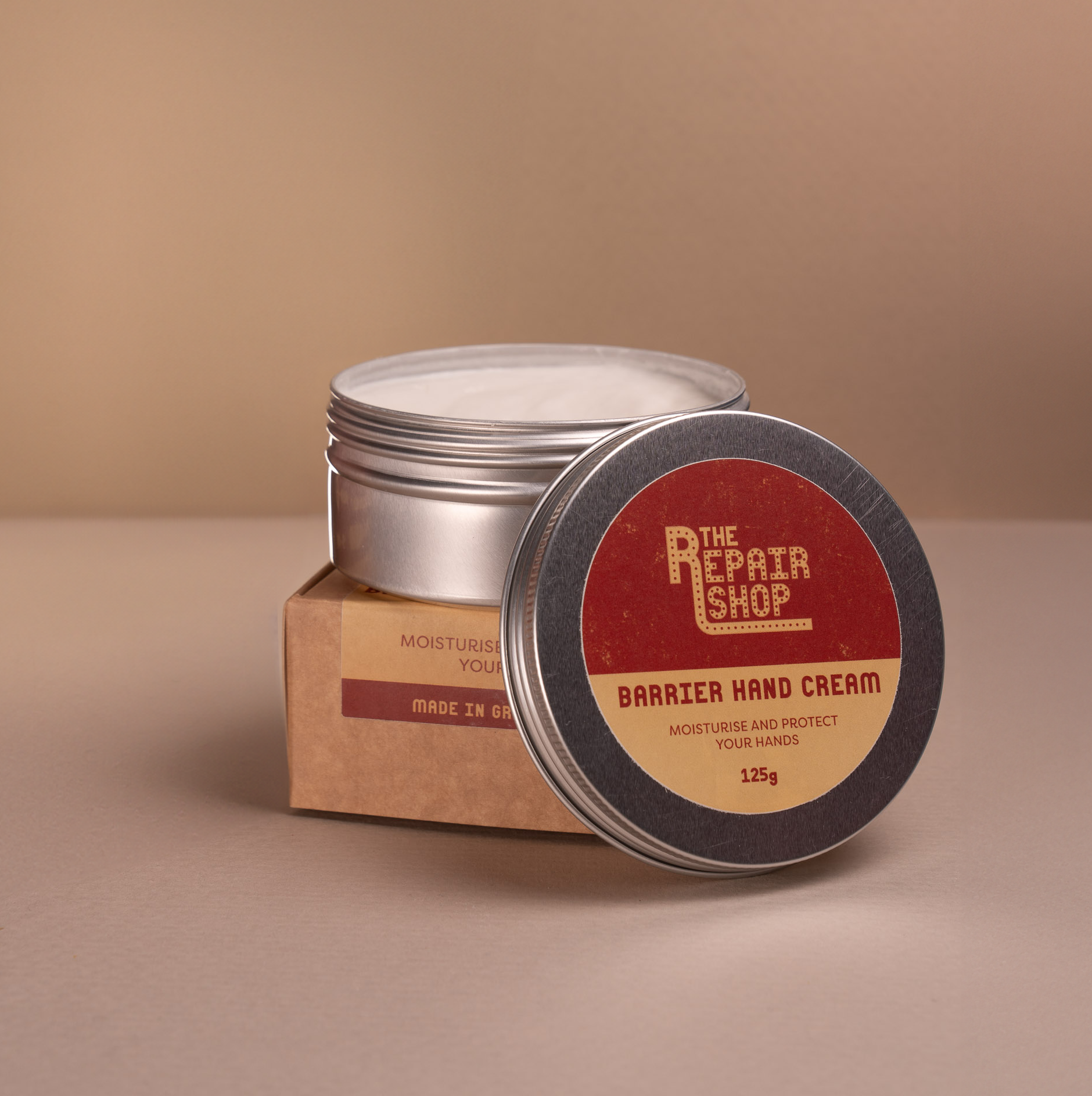 Studio shot of The Repair Shop Barrier Hand Cream. The lid is off showing thick texture of white cream. The aluminium tin sits atop the packaging box. Background is pinky/beige.