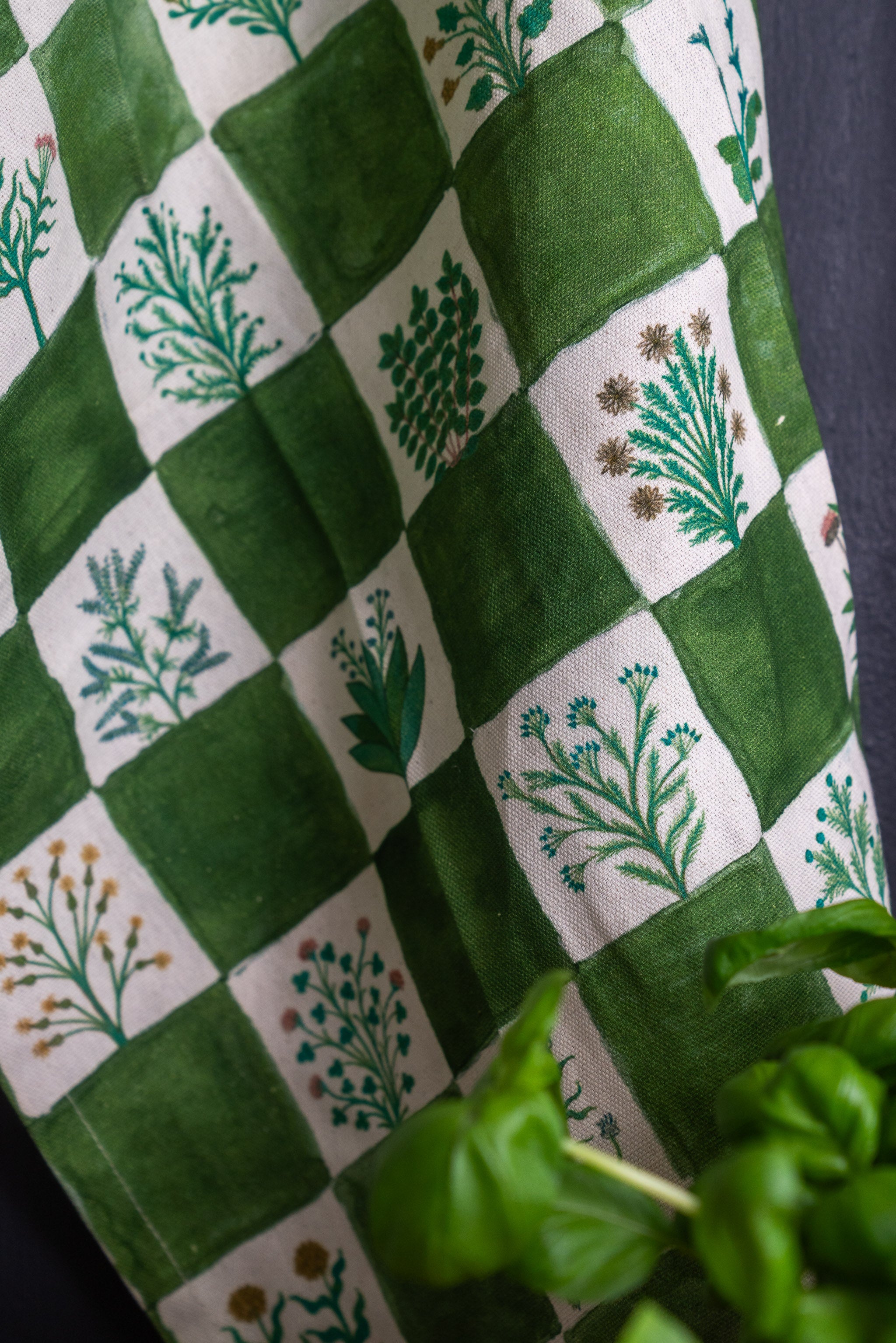 Close up of green and white Herbologist organic cotton tea towel with a basil plant in foreground and a dark blue wall behind.