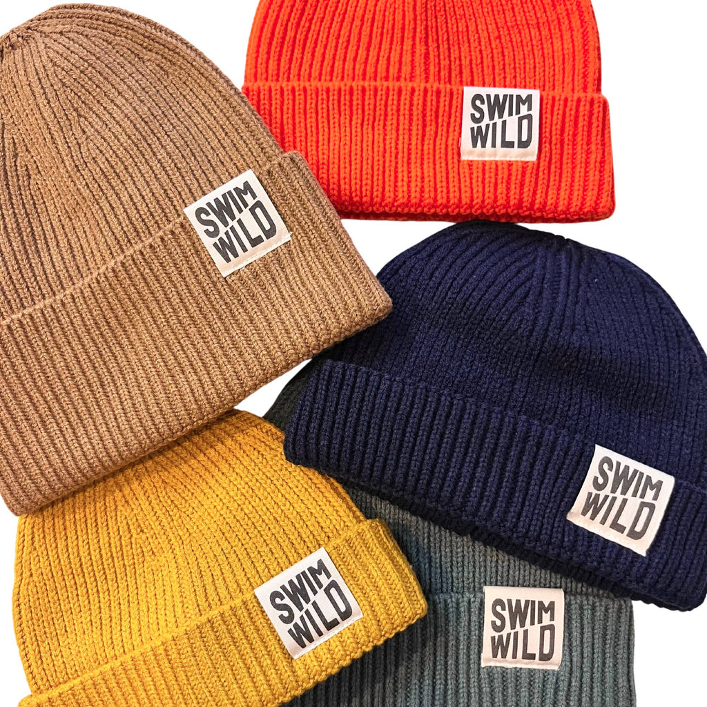 Five knitted fishermans style beanie hats in different colours; bright orange, light brown, navy, mustard yellow and ocean grey. Each hat has a motif reading 'SWIM WILD'. 