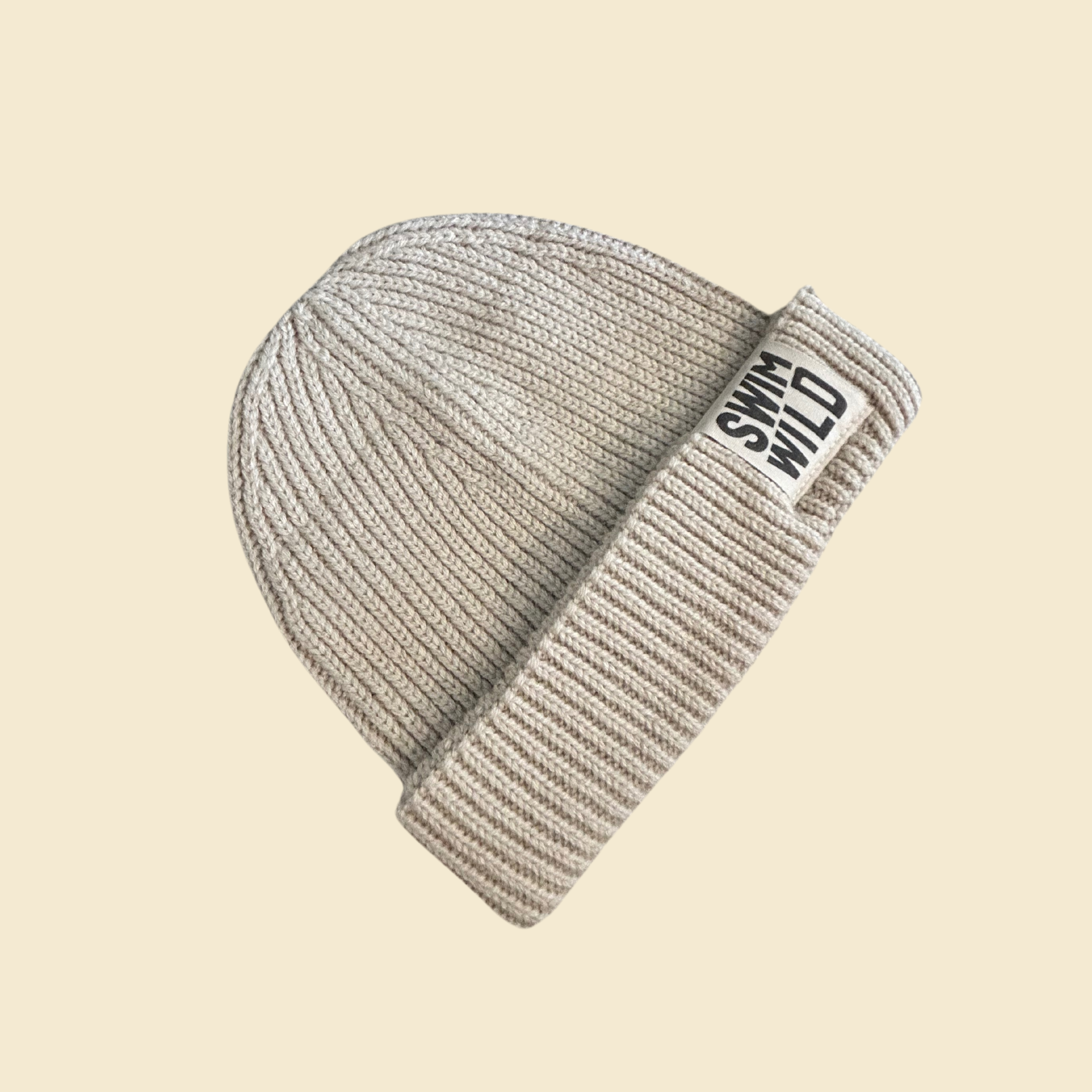 Oat coloured cosy fisherman style beanie hat on cream background. Text on hat reads "SWIM WILD". Great sustainable gift for Wild Swimmers