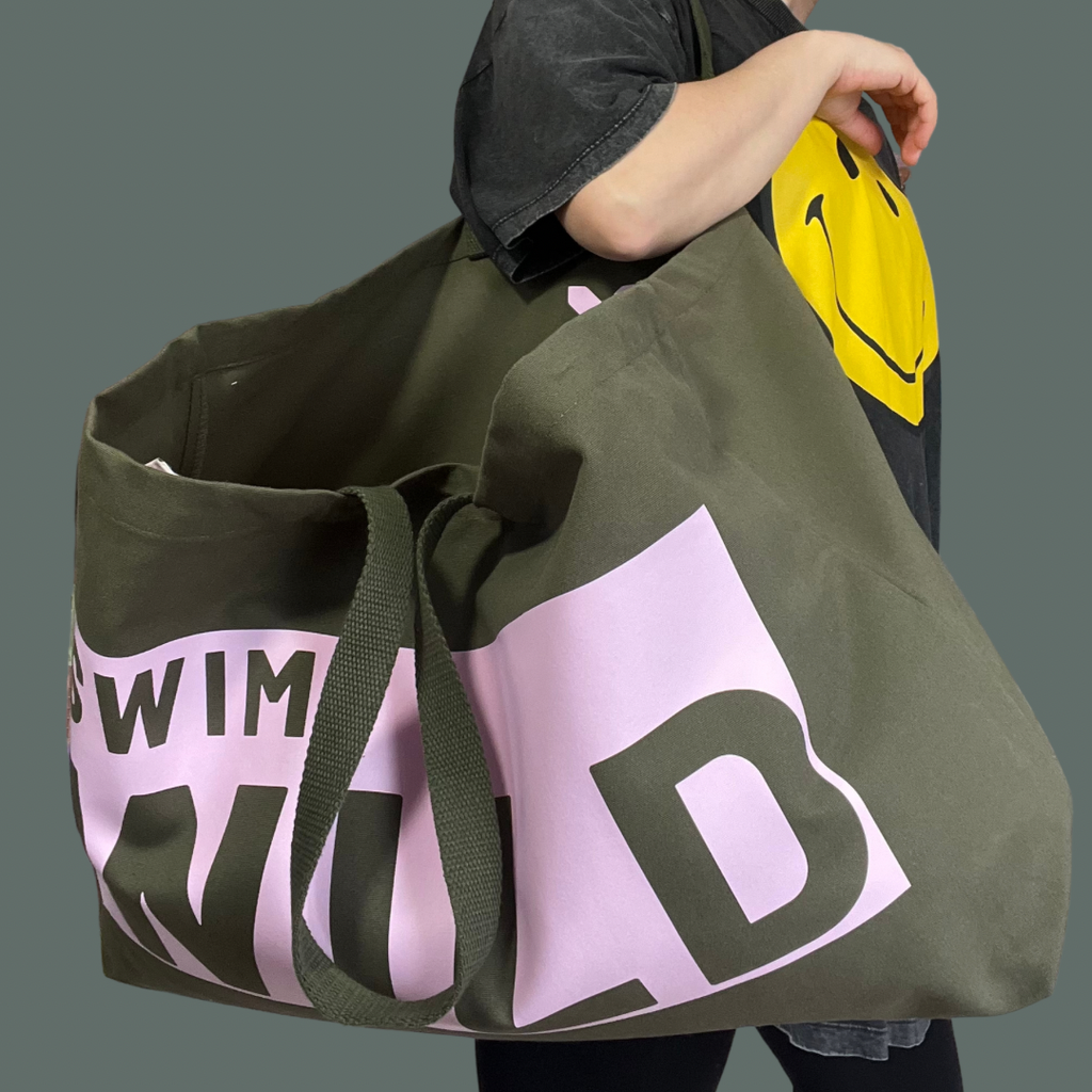 Model wearing jumbo sized olive coloured bag. Text on thick cotton bag is "SWIM WILD" in pink text box Great bag for Wild Swimmers 