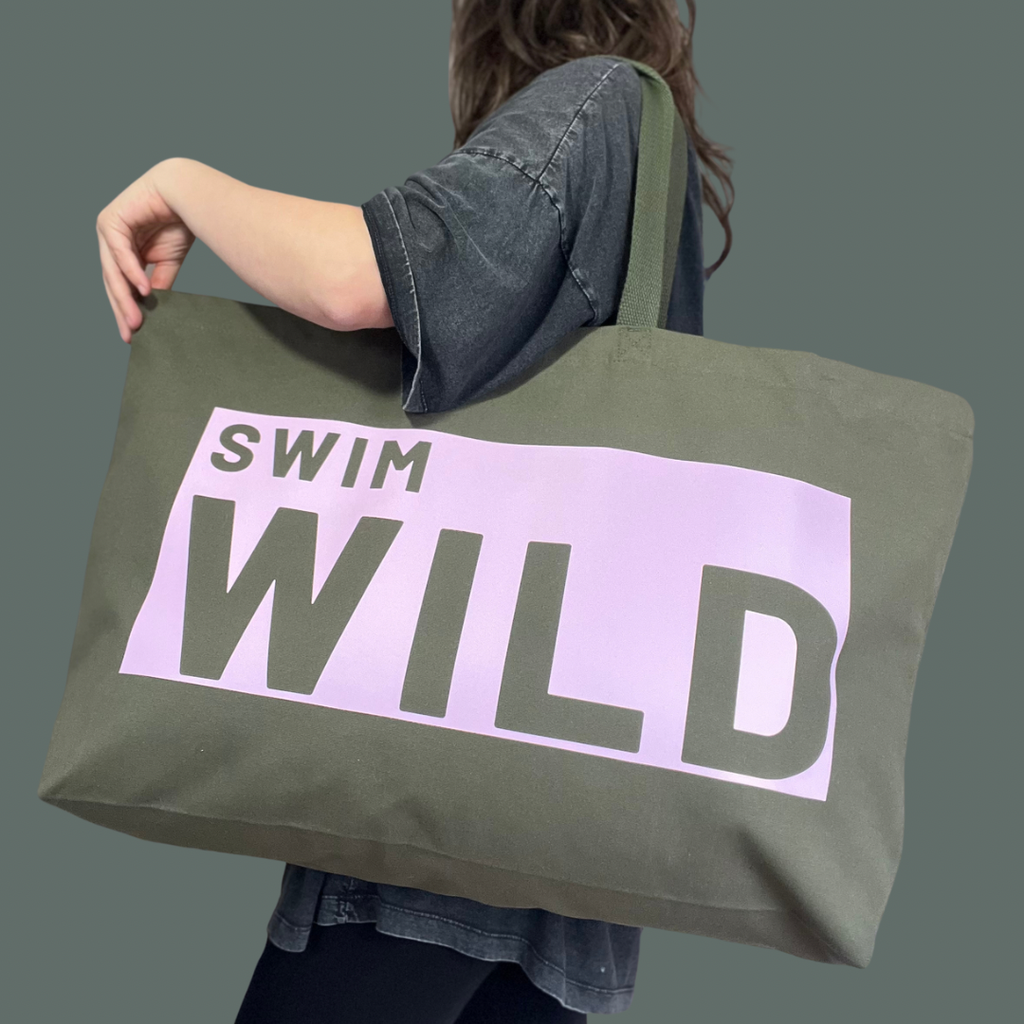 Model wearing jumbo sized olive coloured bag. Text on thick cotton bag is "SWIM WILD" in pale pink text box. Great gift for Wild Swimmers 