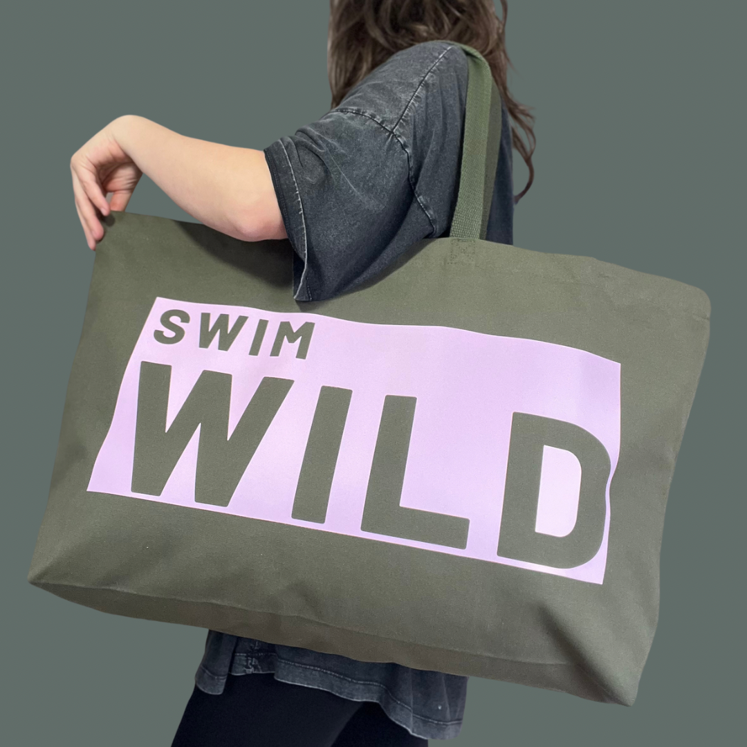 Model wearing jumbo sized olive coloured bag. Text on thick cotton bag is "SWIM WILD" in pale pink text box. Great gift for Wild Swimmers 