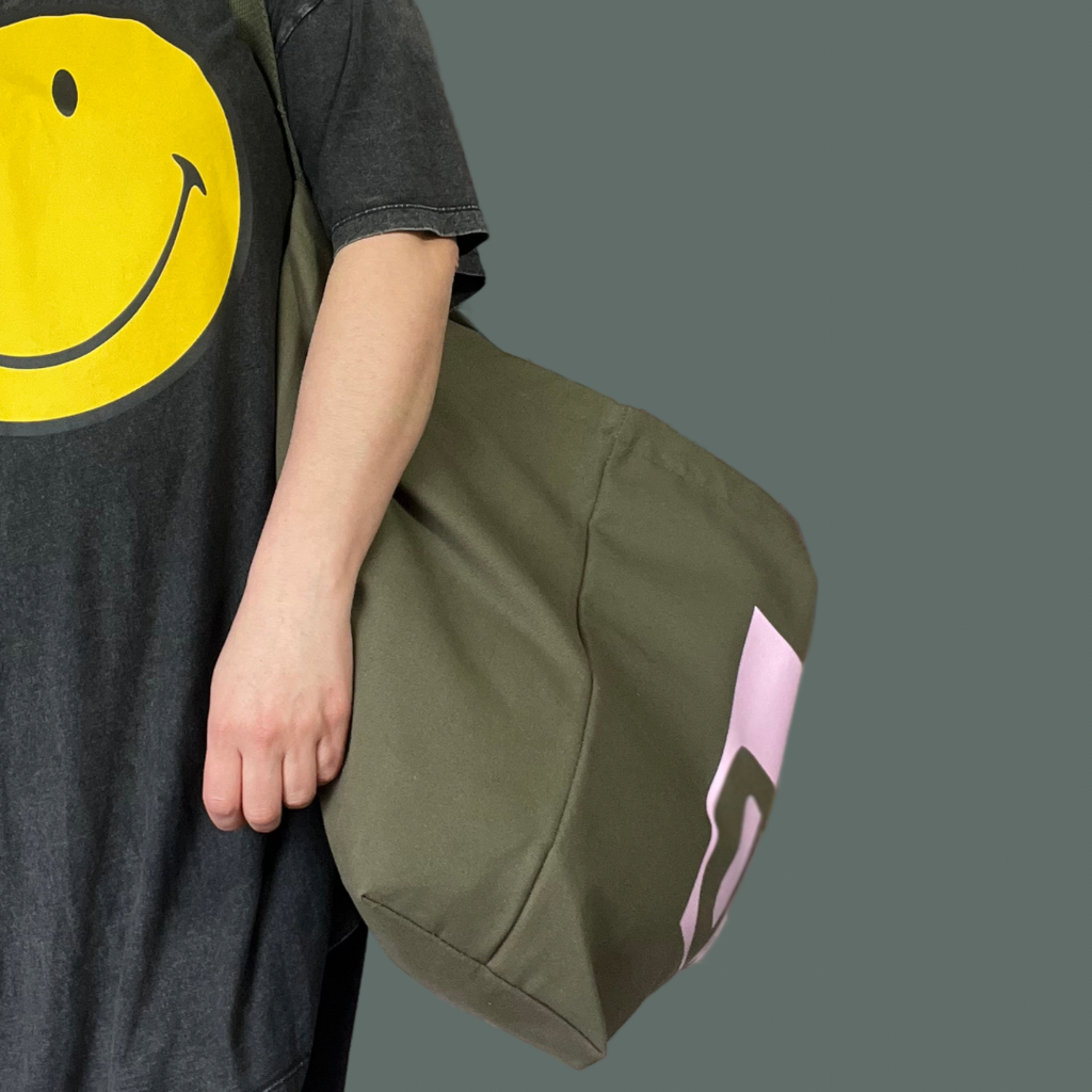 Olive green jumbo SWIM WILD bag viewed side on. Pale pink text box partially visible. Showing width of bag. Model is seen from neck down in a large grey t shirt with yellow smiley face. SWIM WILD bag for Wild Swimmers